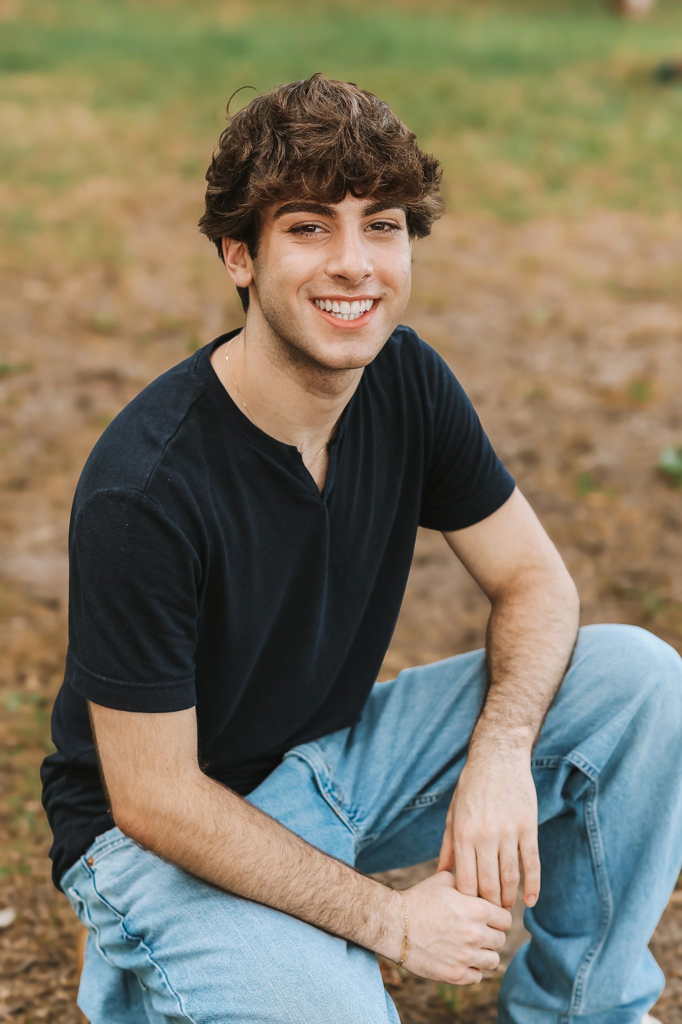 Happy young man in black t-shirt and light jeans sitting outdoors smiling in natural setting.