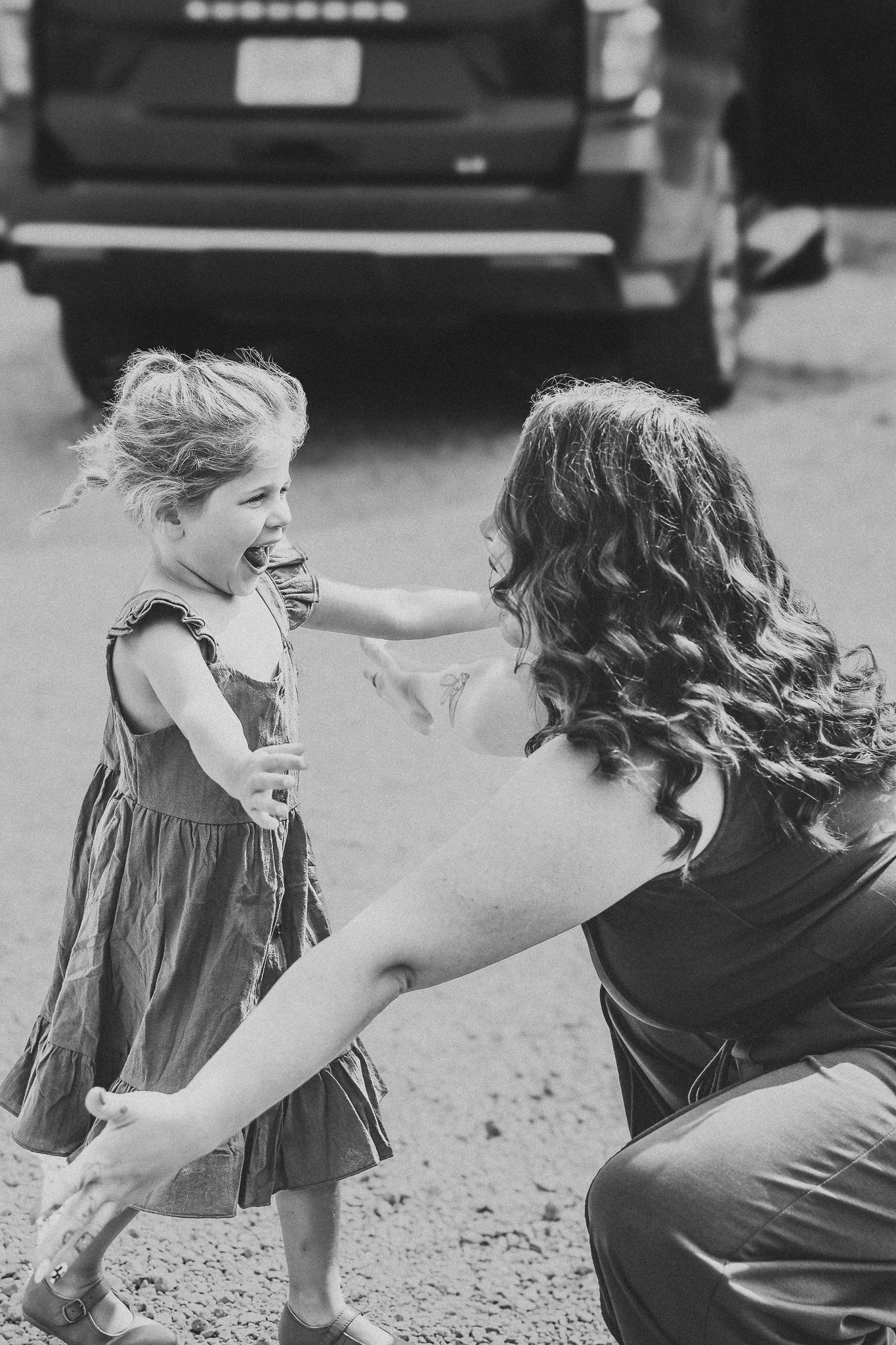 Black and white photo of joyful little girl reaching toward mother crouching in parking lot.