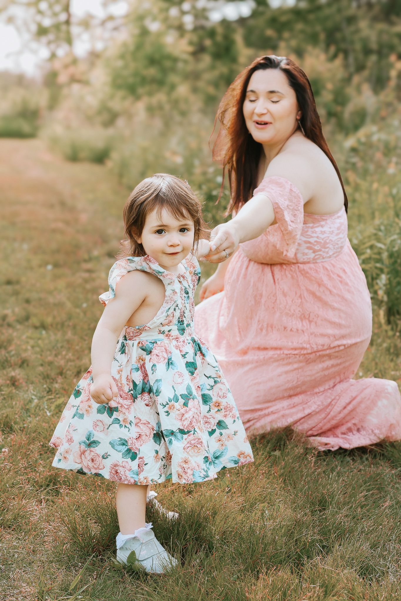 Pregnant woman in pink lace gown holds toddler girl's hand in a garden setting.
