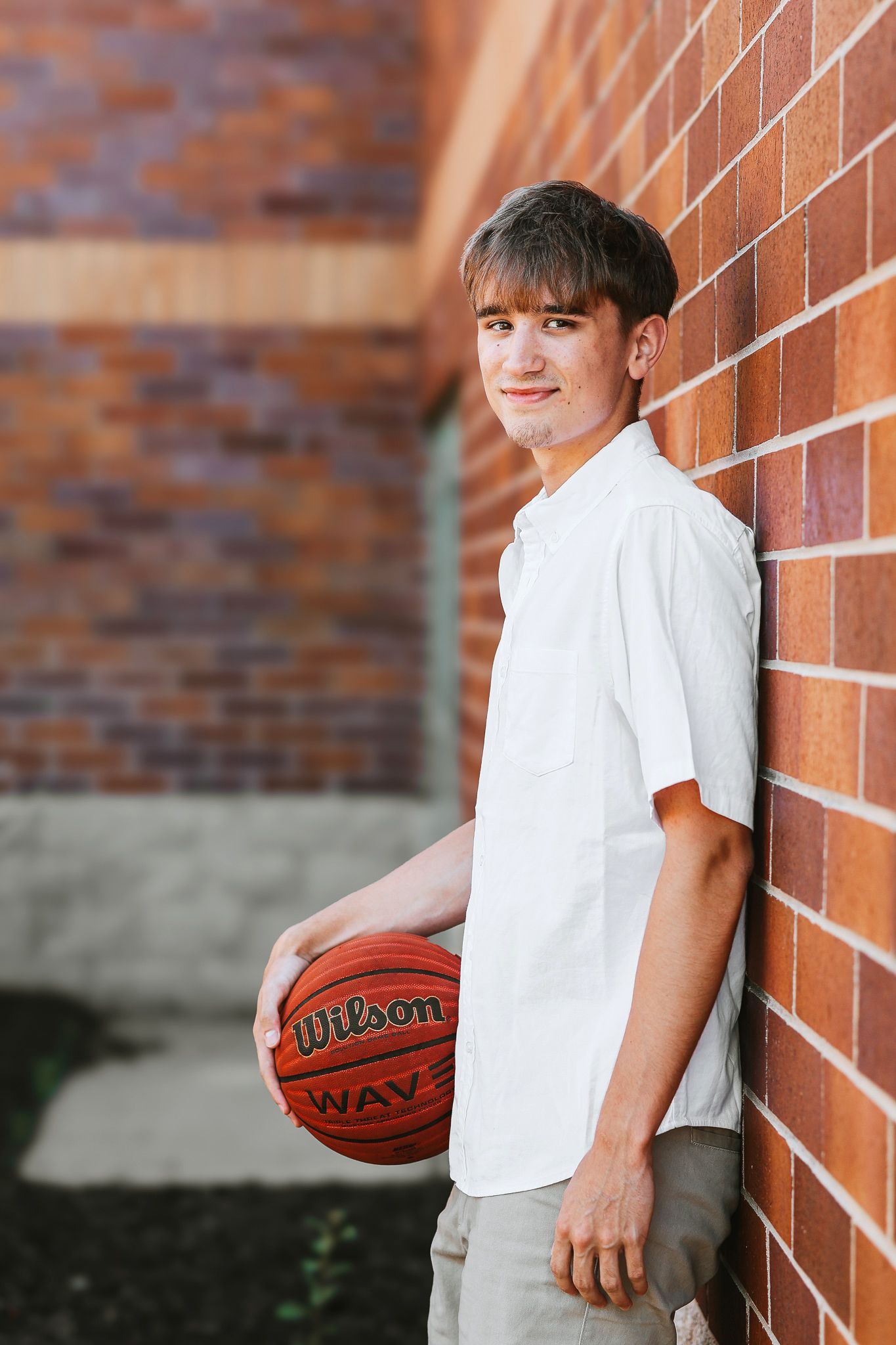 Teen boy in white shirt leaning against red brick wall holding a Wilson basketball outdoors.