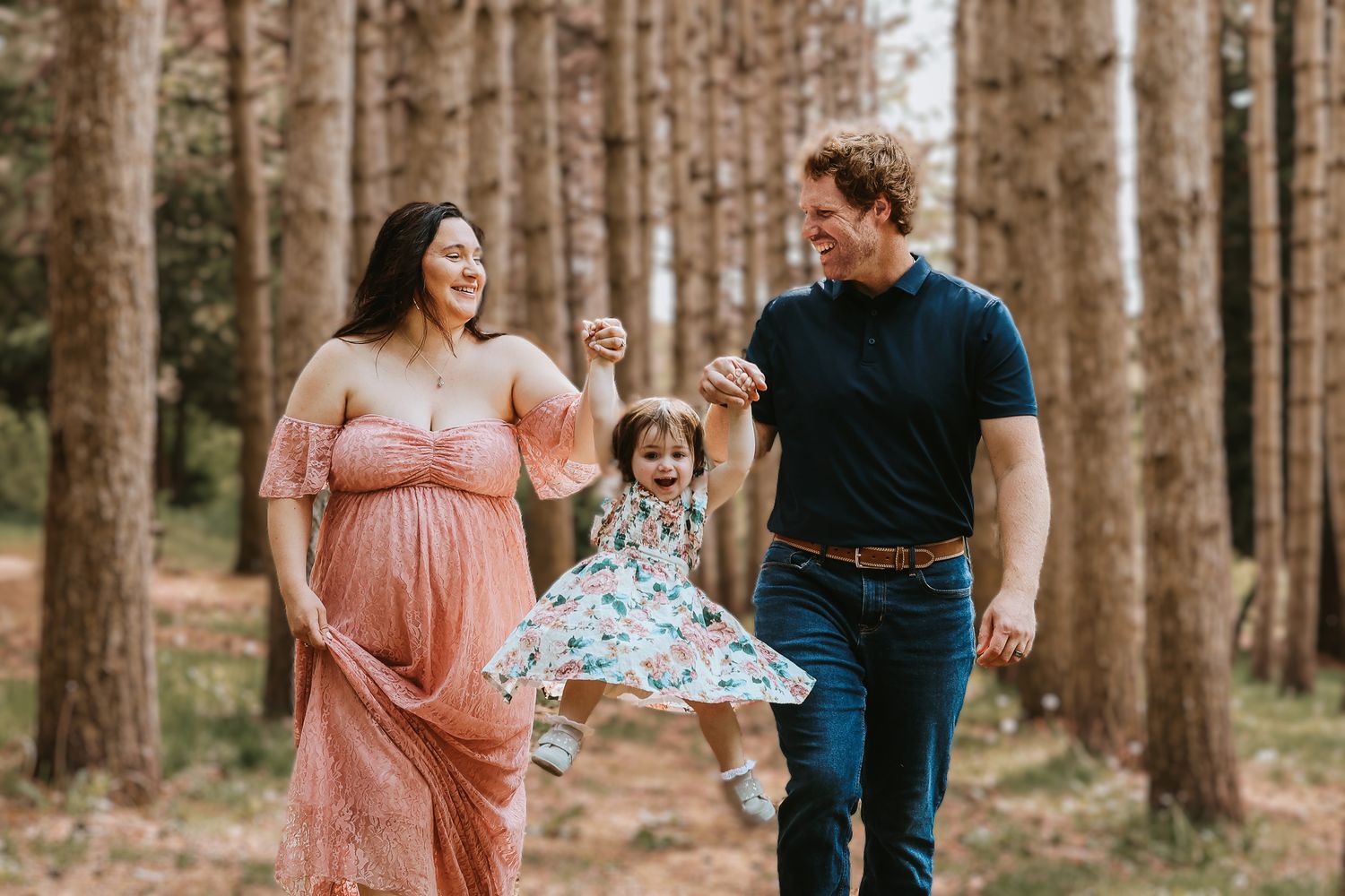 Joyful family swinging toddler between parents walking through a pine forest during maternity session.