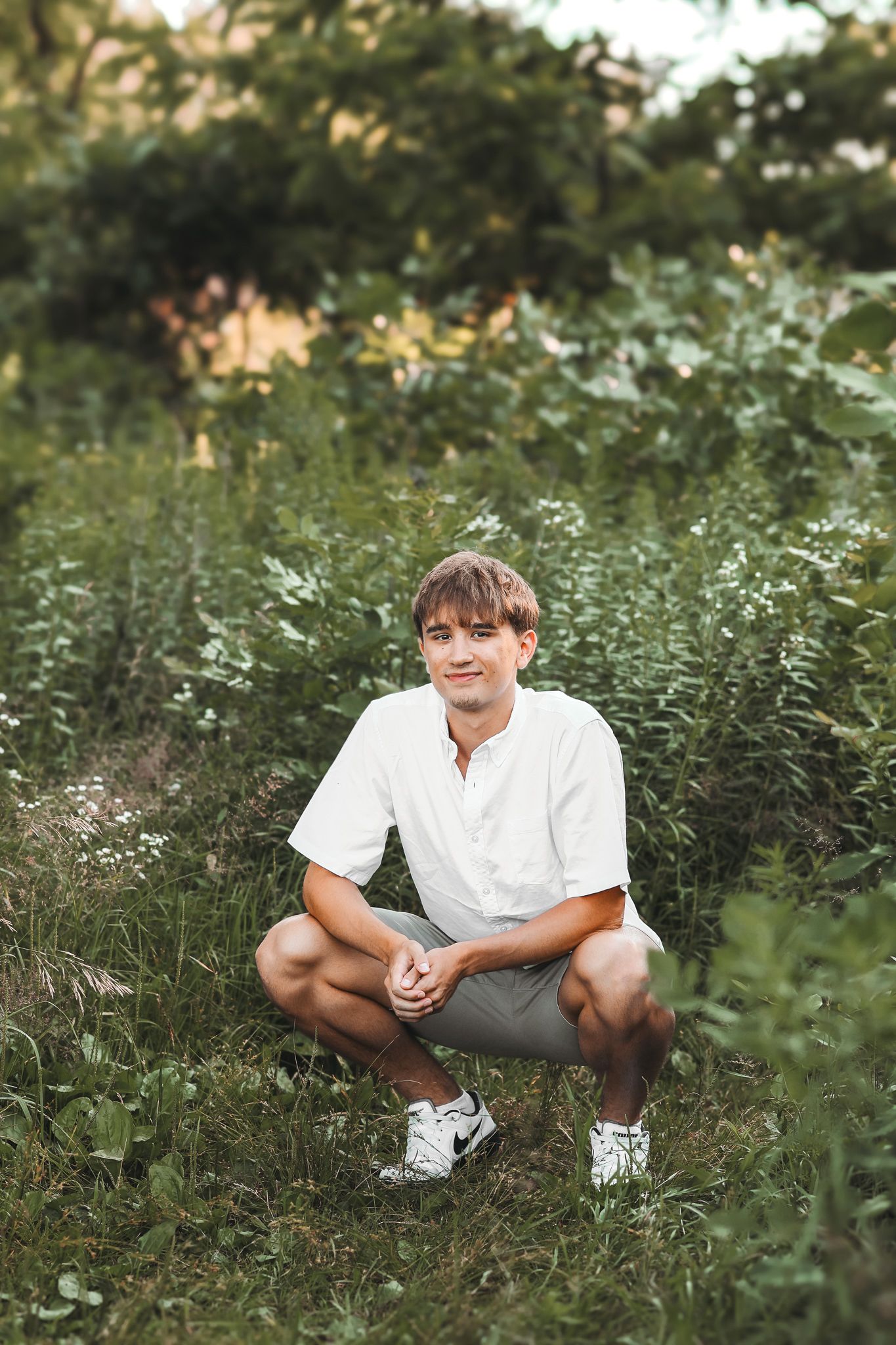 Teen boy in white shirt and gray shorts crouching in a lush green outdoor setting.