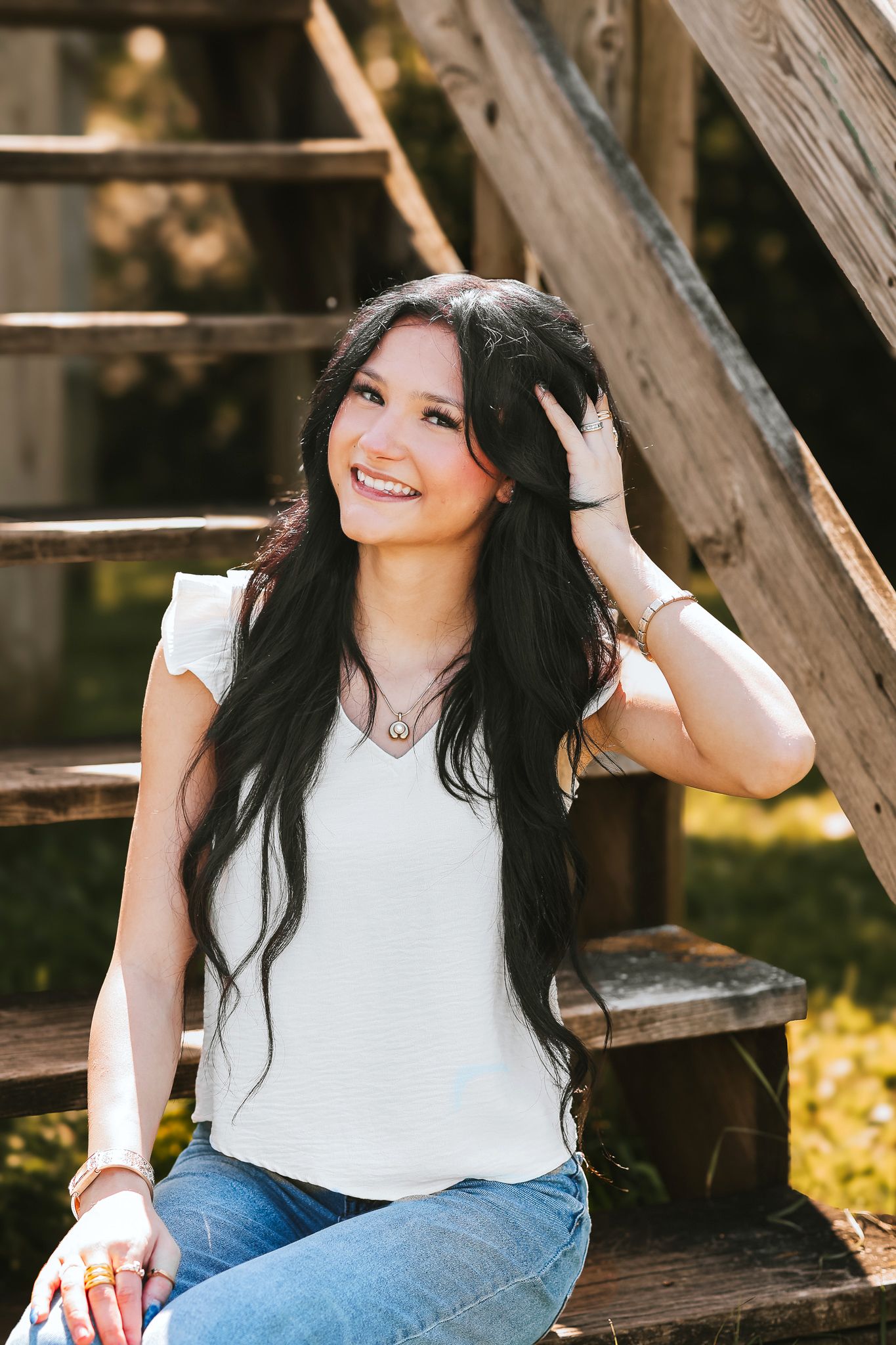 Smiling teenage girl with long black hair sitting on wooden steps in jeans and a white top on a sunny day.