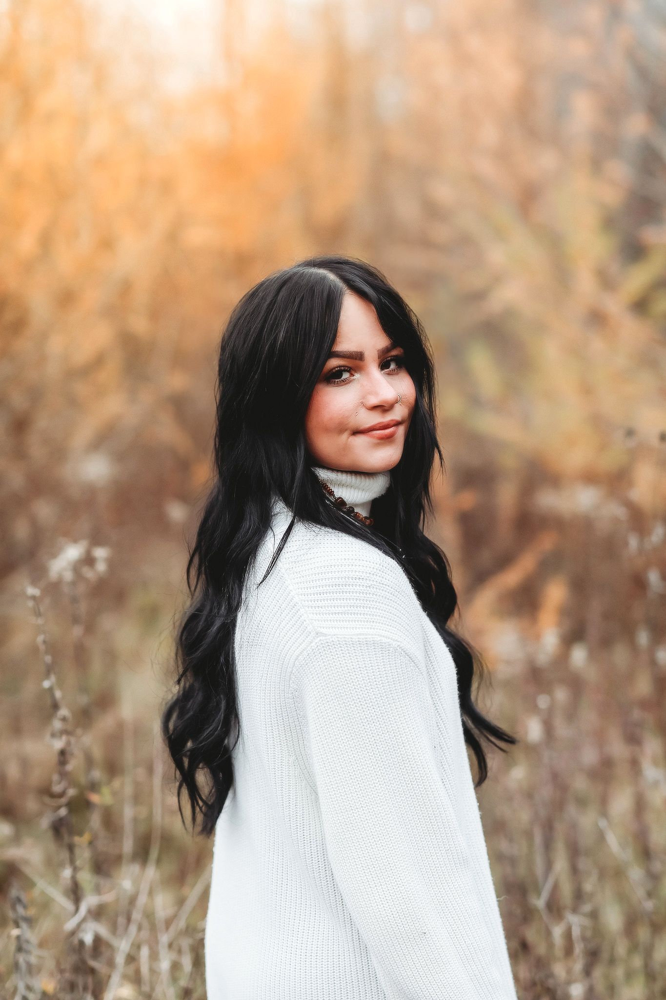 Young woman with long black hair in white turtleneck sweater smiling outdoors in autumn field.