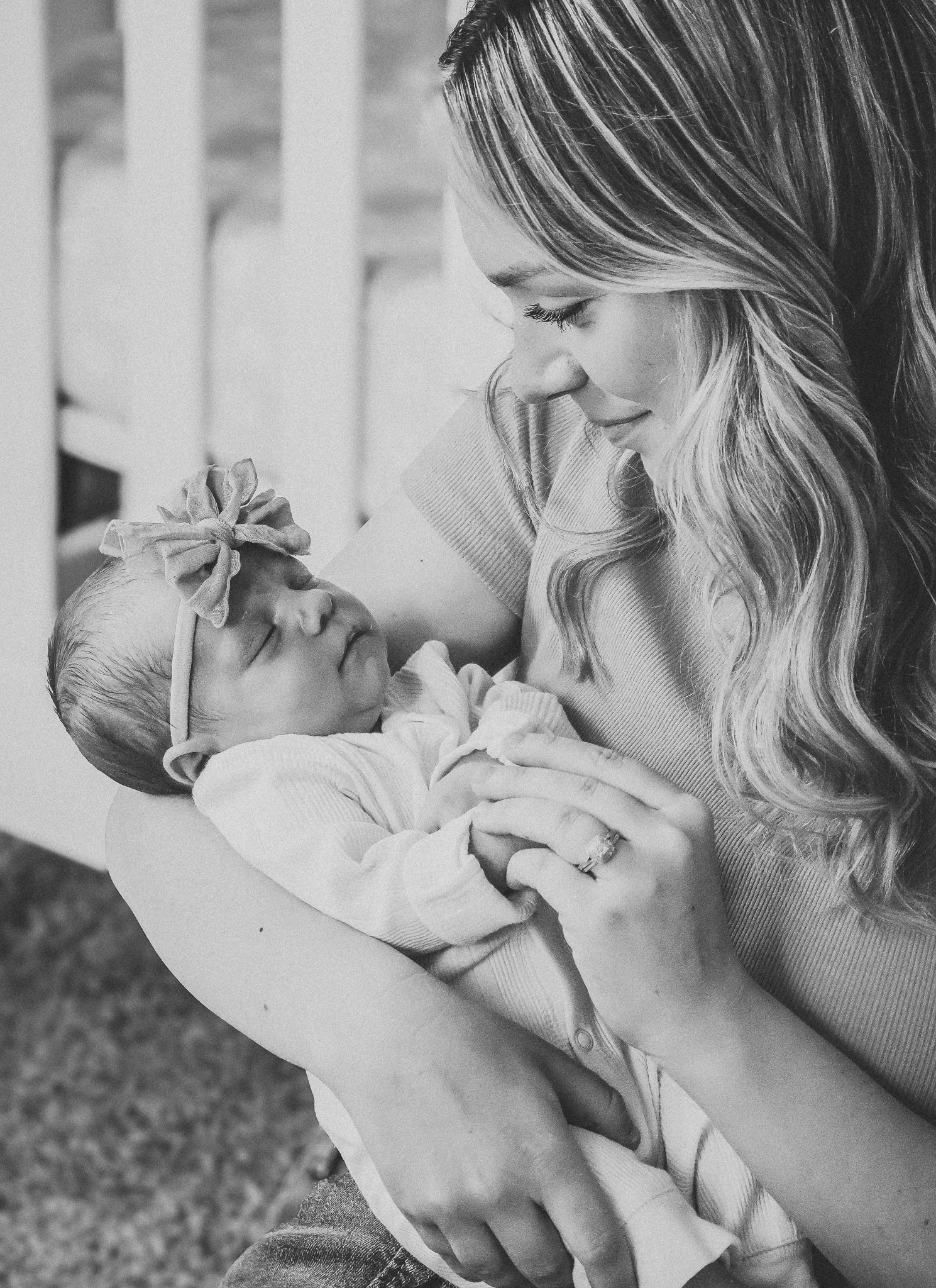 Black and white photo of a smiling mother tenderly holding her newborn baby girl with a bow headband.