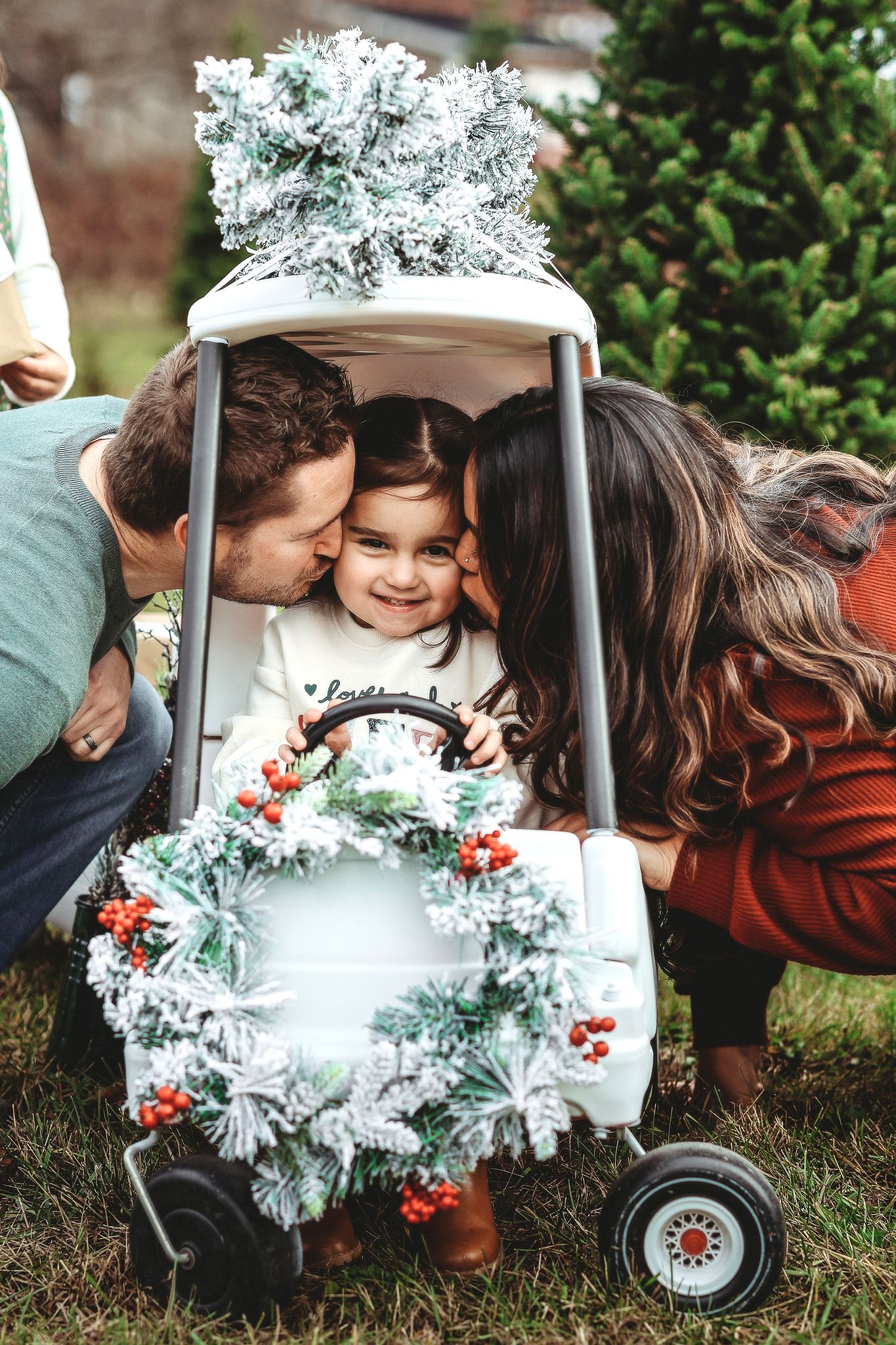 Parents kissing smiling toddler in a decorated toy car with Christmas wreath and tree.