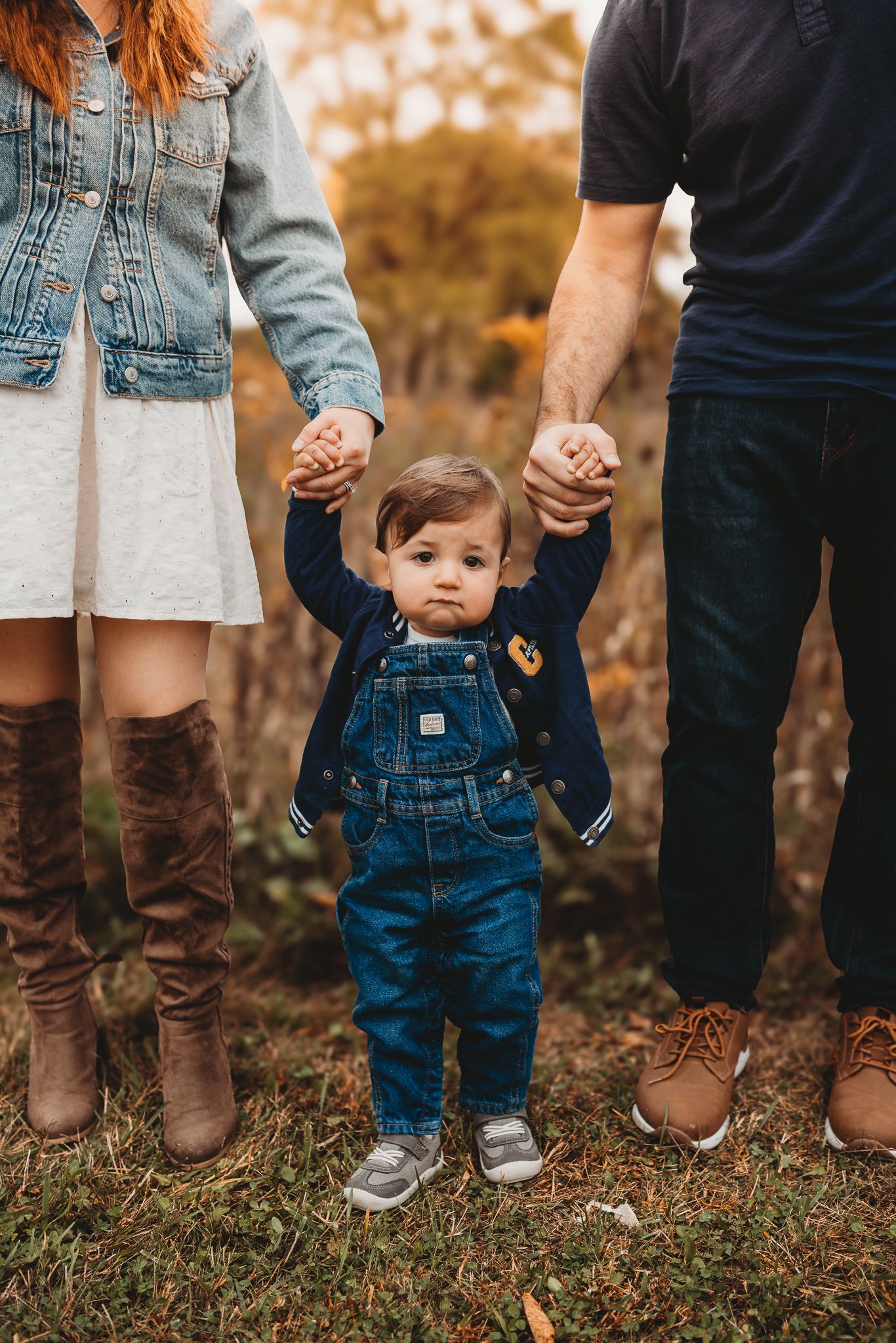 Toddler in denim overalls holds parents hands while standing in a fall outdoor landscape.