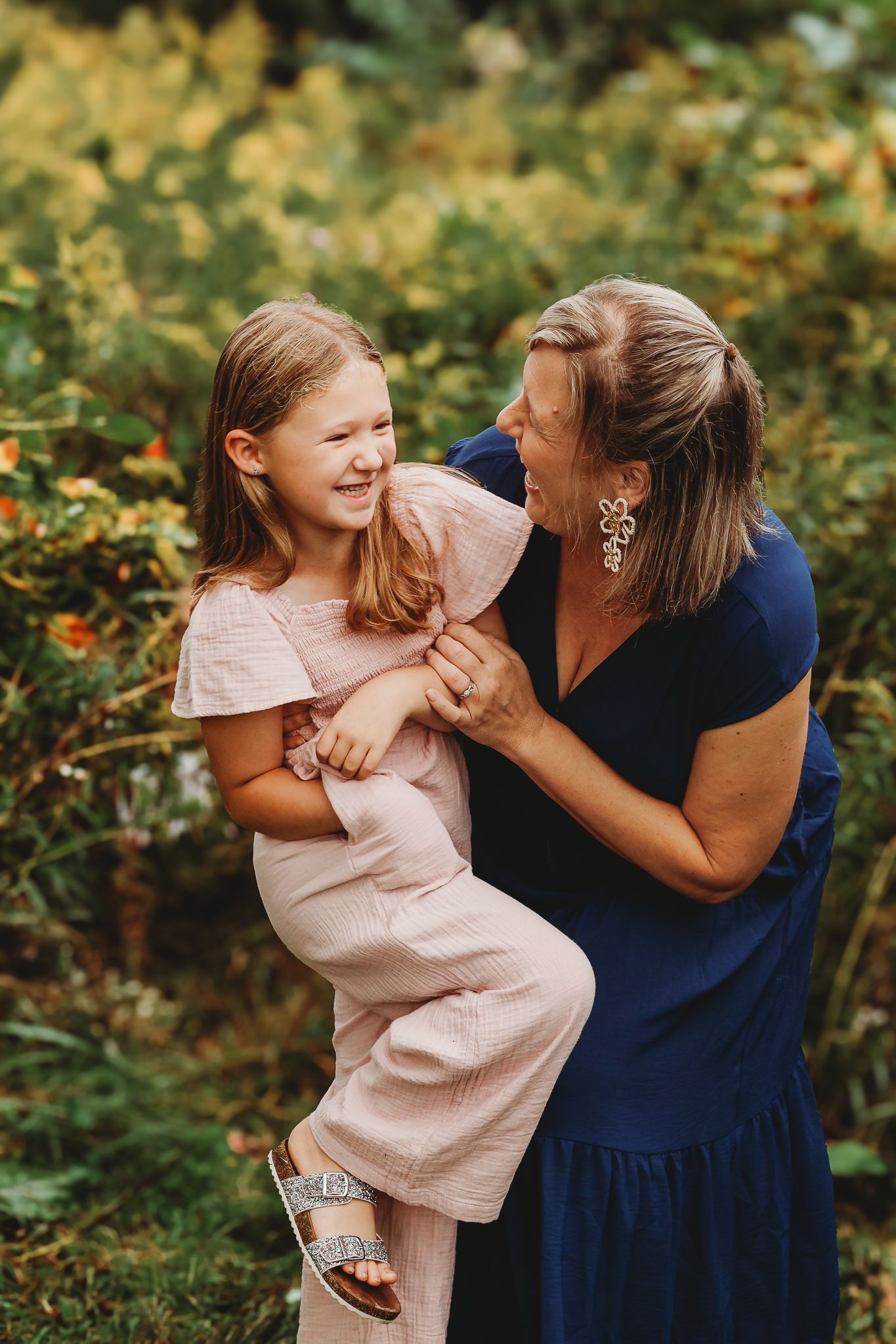A mother laughs while holding her smiling daughter outdoors among lush green and yellow foliage.