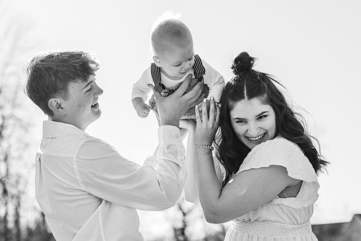 Black and white photo of joyful young parents lifting laughing baby in sunny outdoor setting.