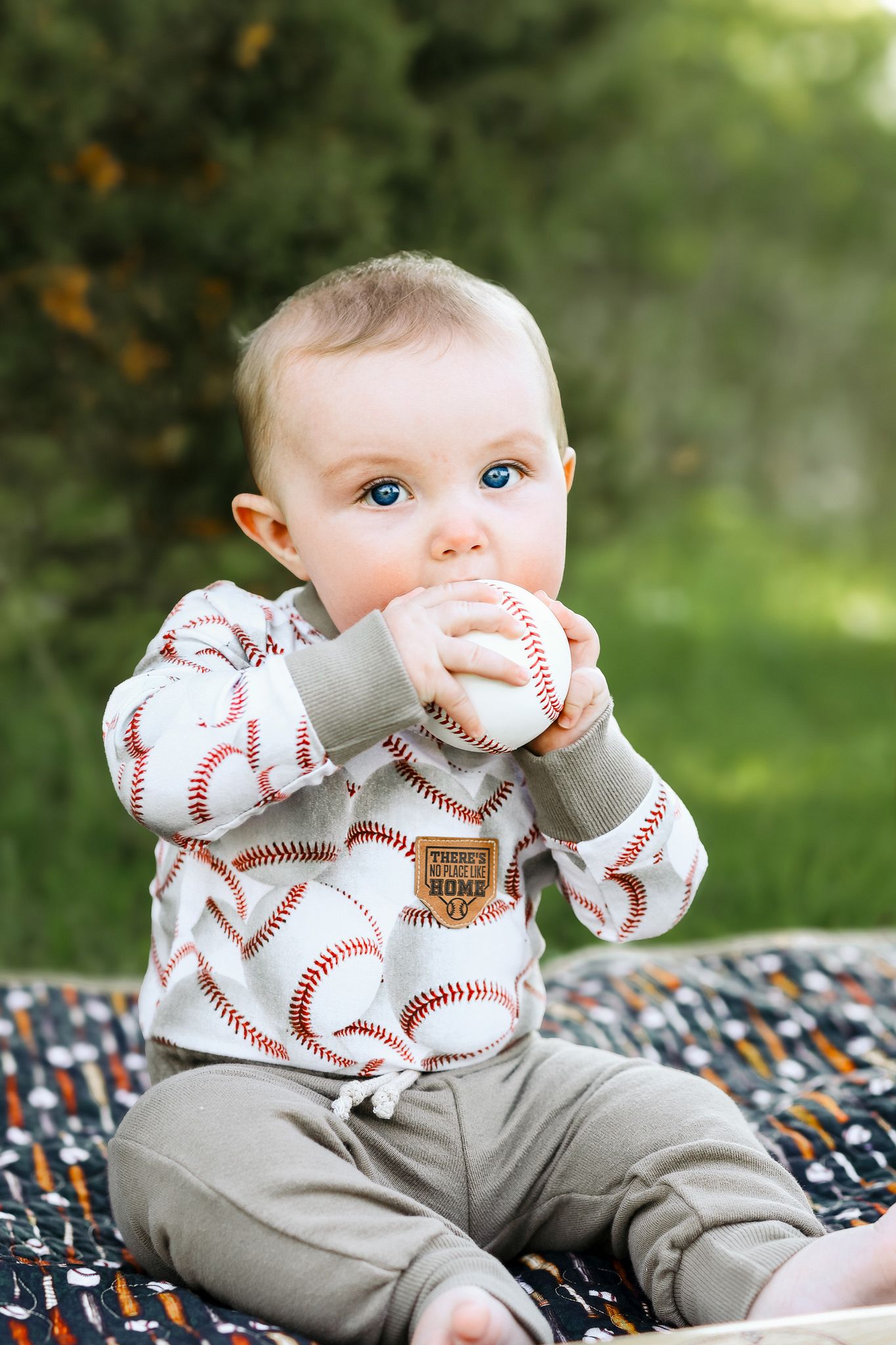 Baby in baseball-print outfit chewing on a baseball while sitting outdoors on a blanket.