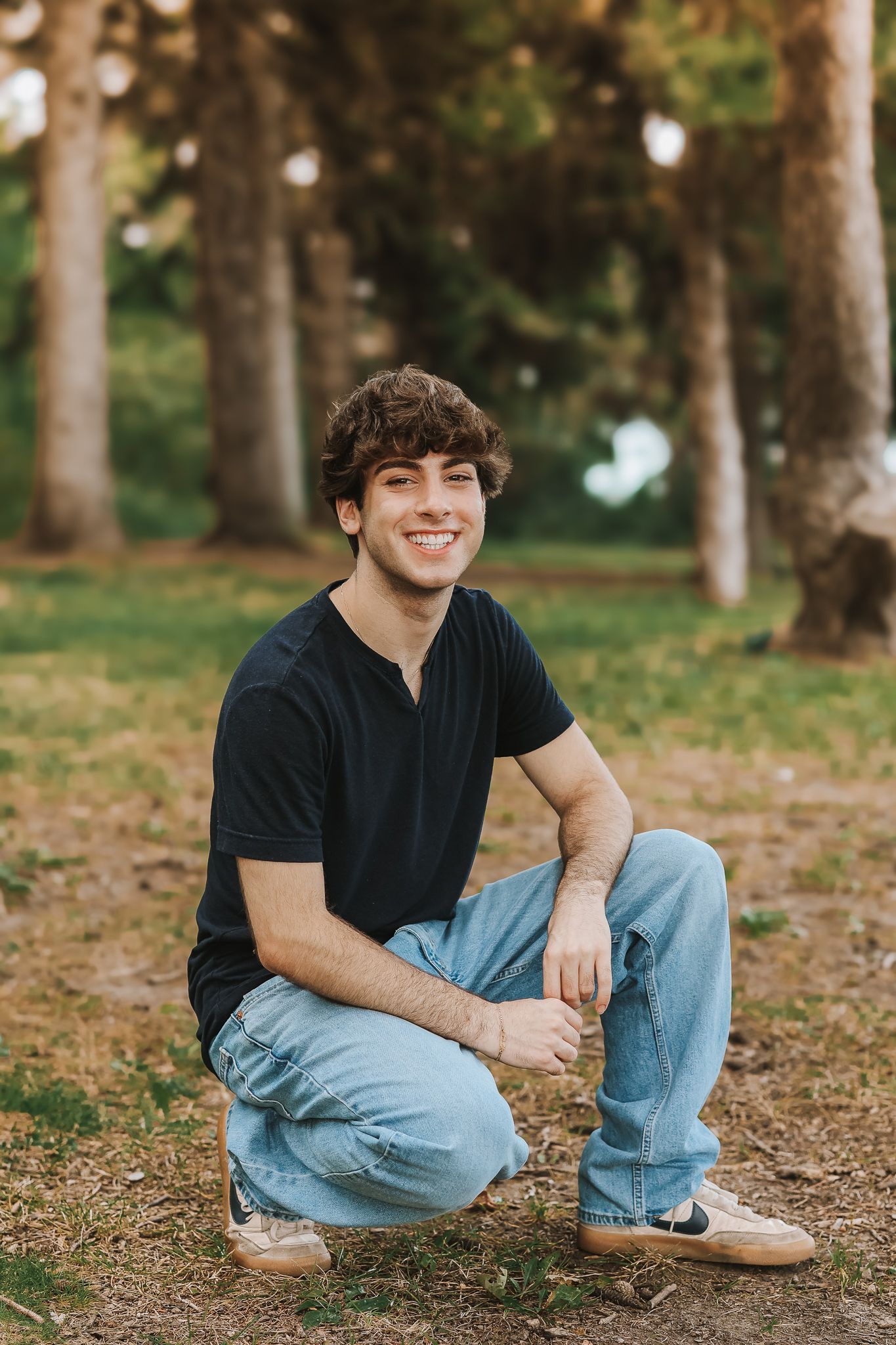 Smiling teenage boy in black shirt and jeans crouching on grass in front of pine trees.