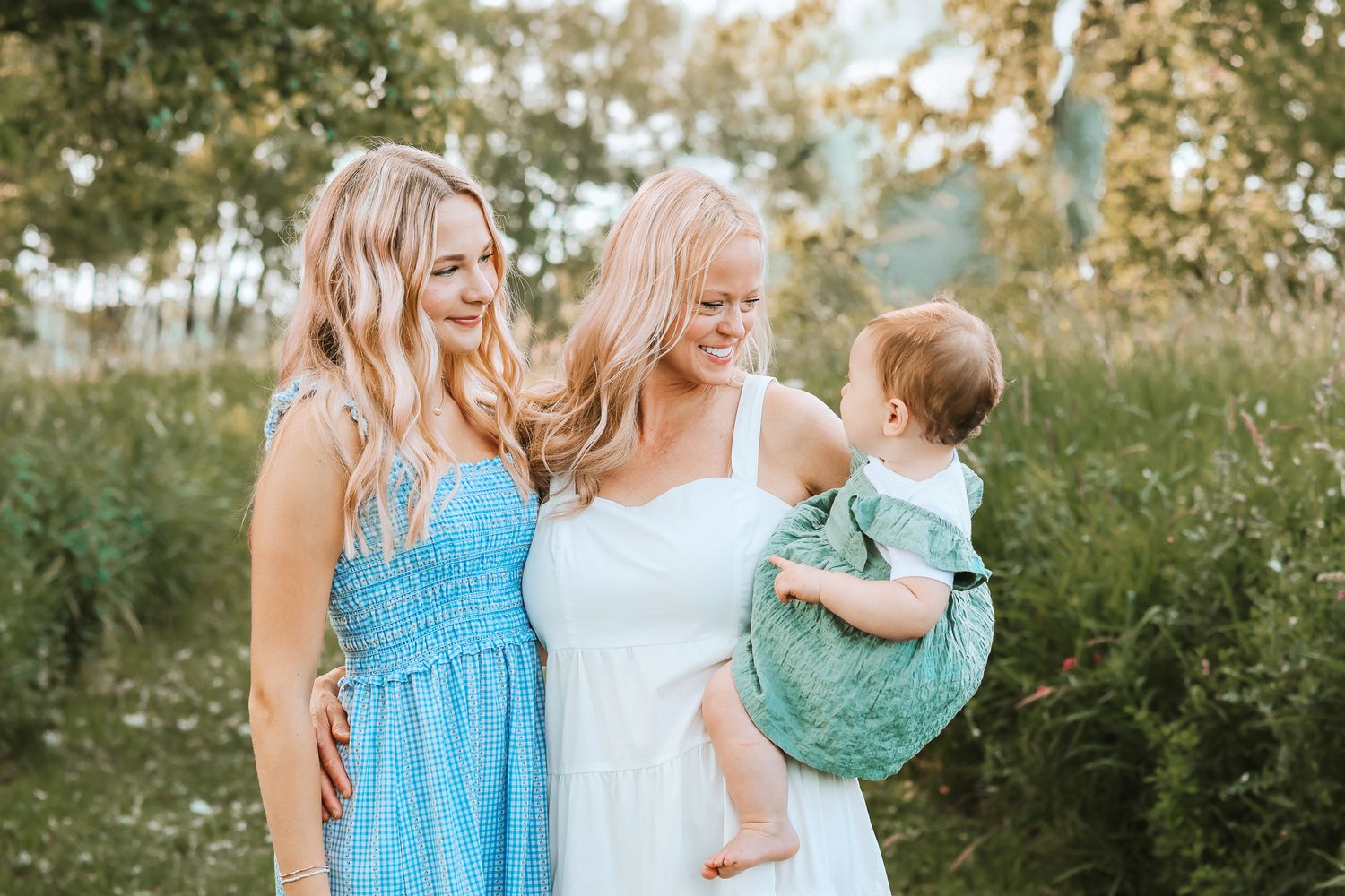 Two blonde women in blue and white dresses smiling at an adorable baby in a green outfit outdoors.