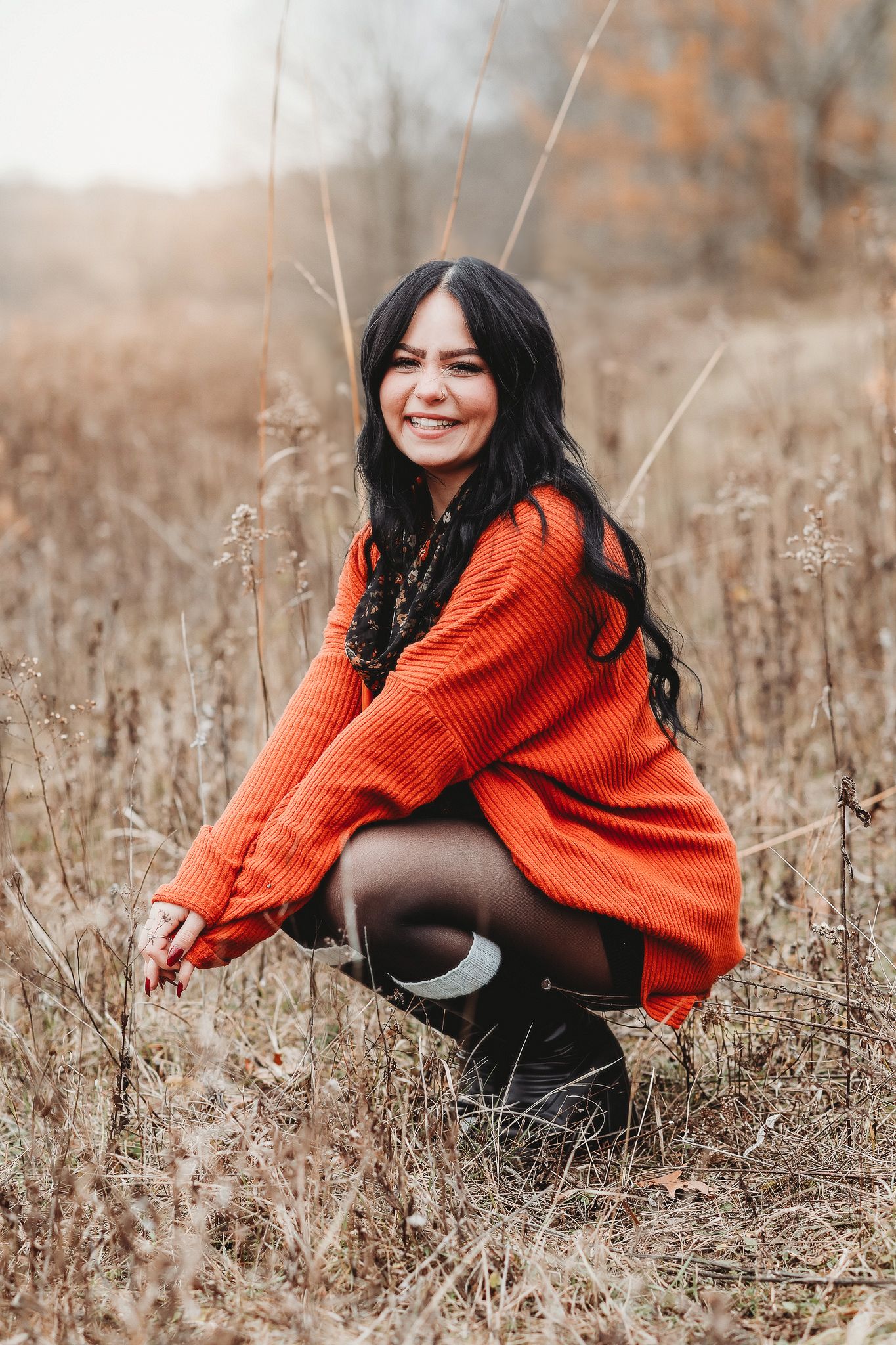 Smiling young woman in orange sweater crouching in a dry autumn field with tall brown grass.