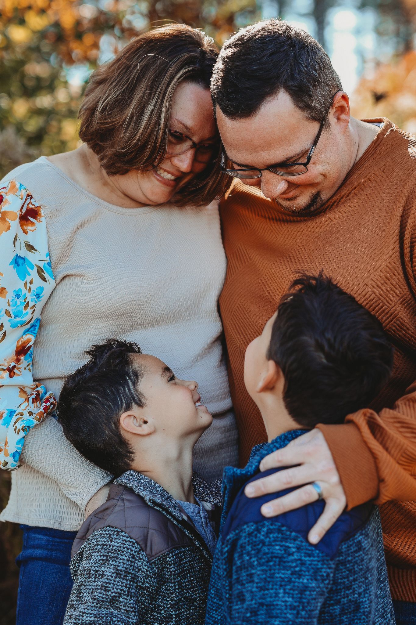 Family of four sharing a warm group hug outdoors in autumn, two boys looking up at parents.