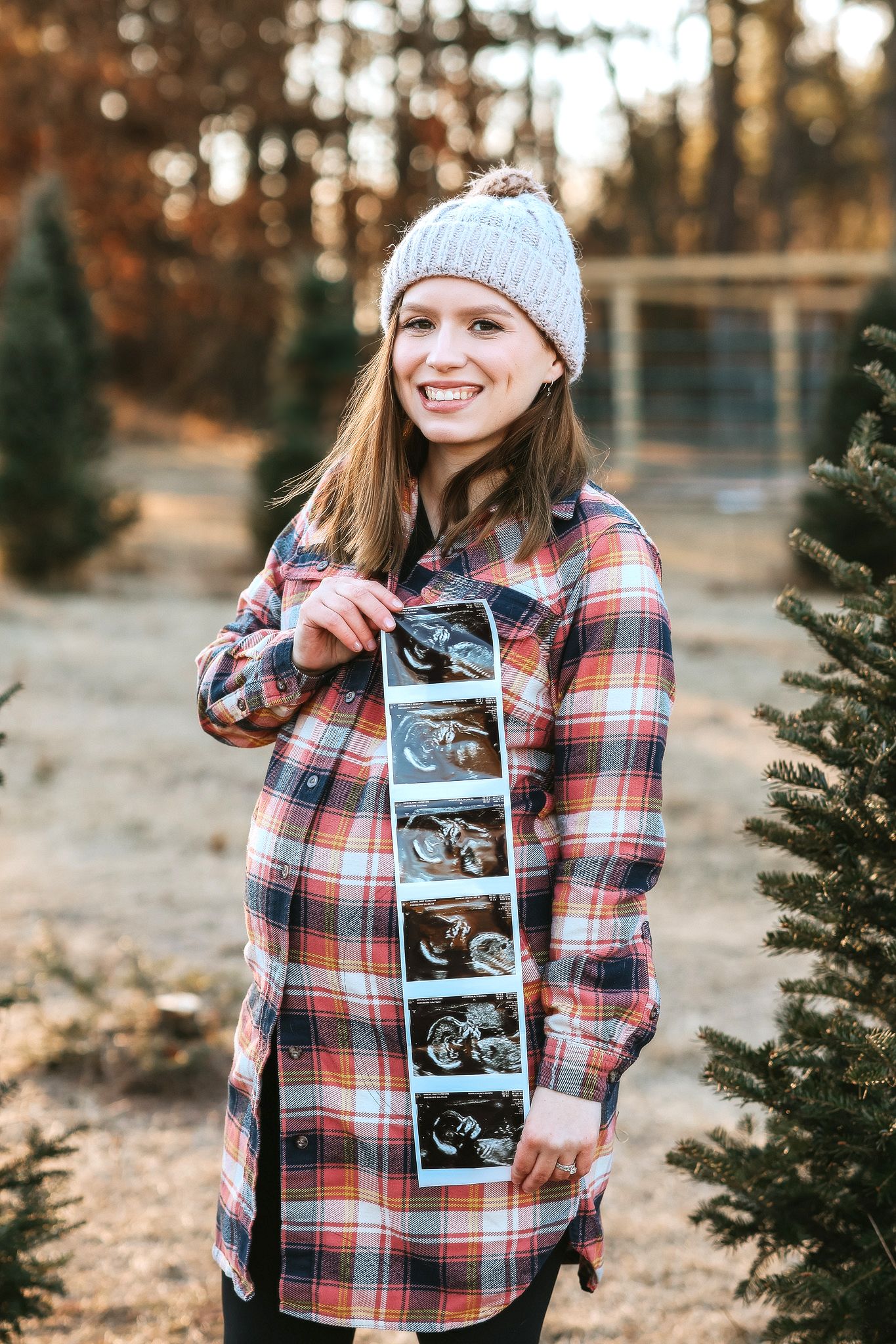 Smiling pregnant woman in gray beanie and plaid shirt holding ultrasound photos near pine trees.