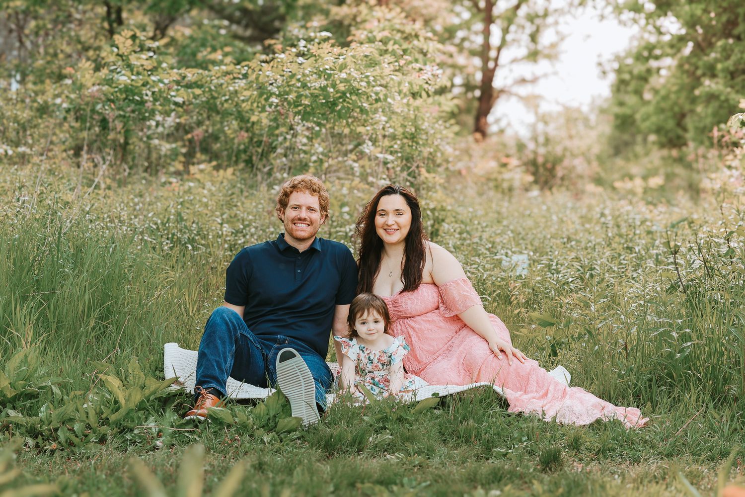 Happy family of three sitting together in a lush green meadow during maternity session.