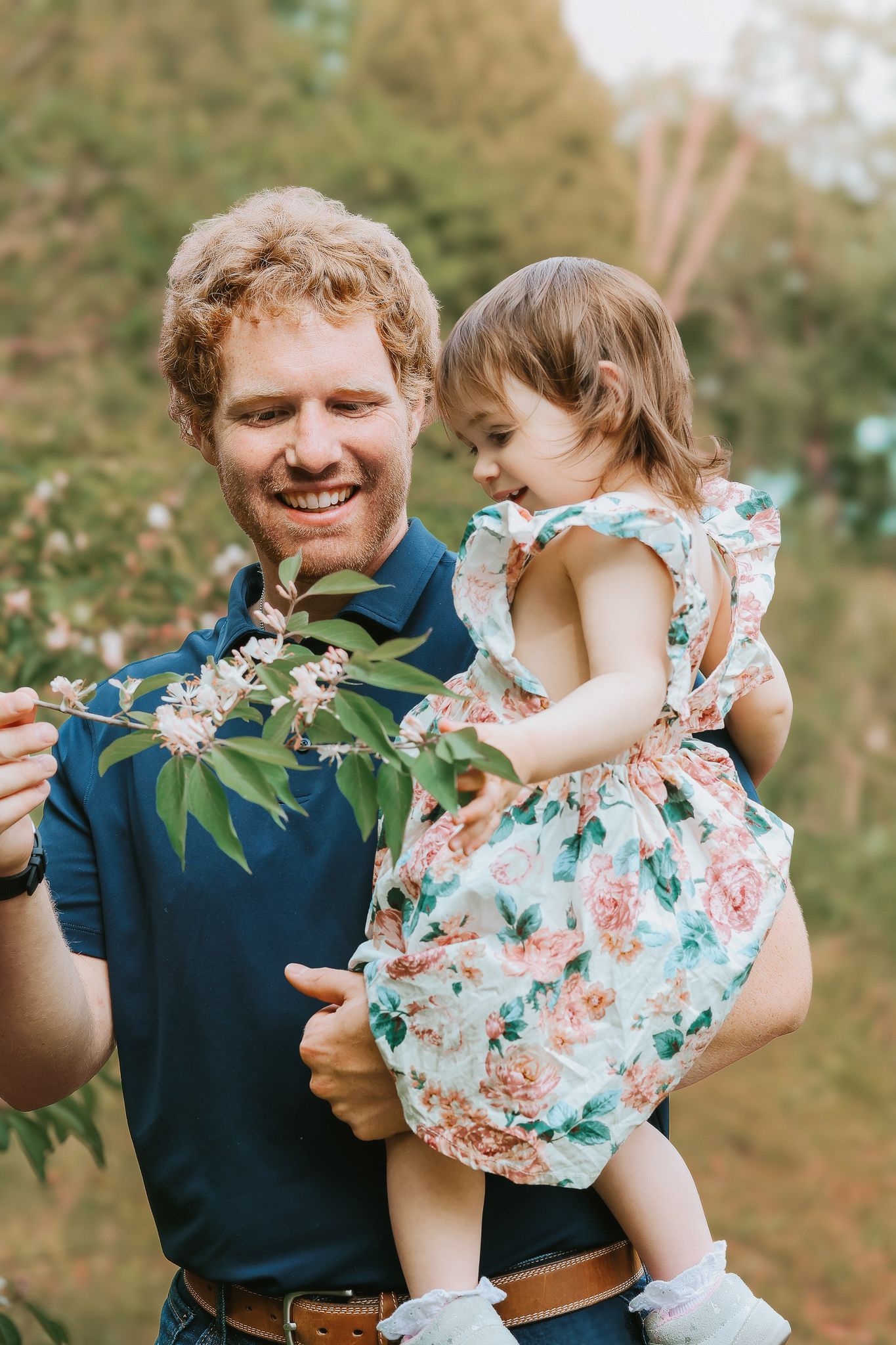 Smiling redhead father holding toddler daughter in floral dress while showing her flowers on a branch outdoors.
