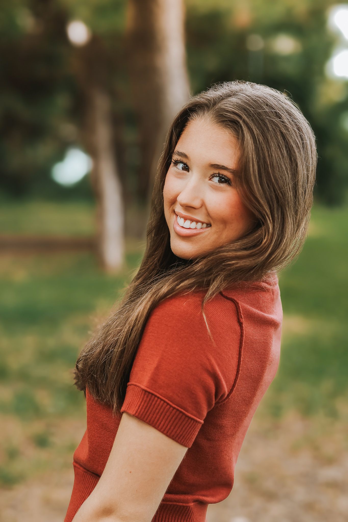 Teenage girl in rust red top smiling over shoulder during outdoor senior portrait session.