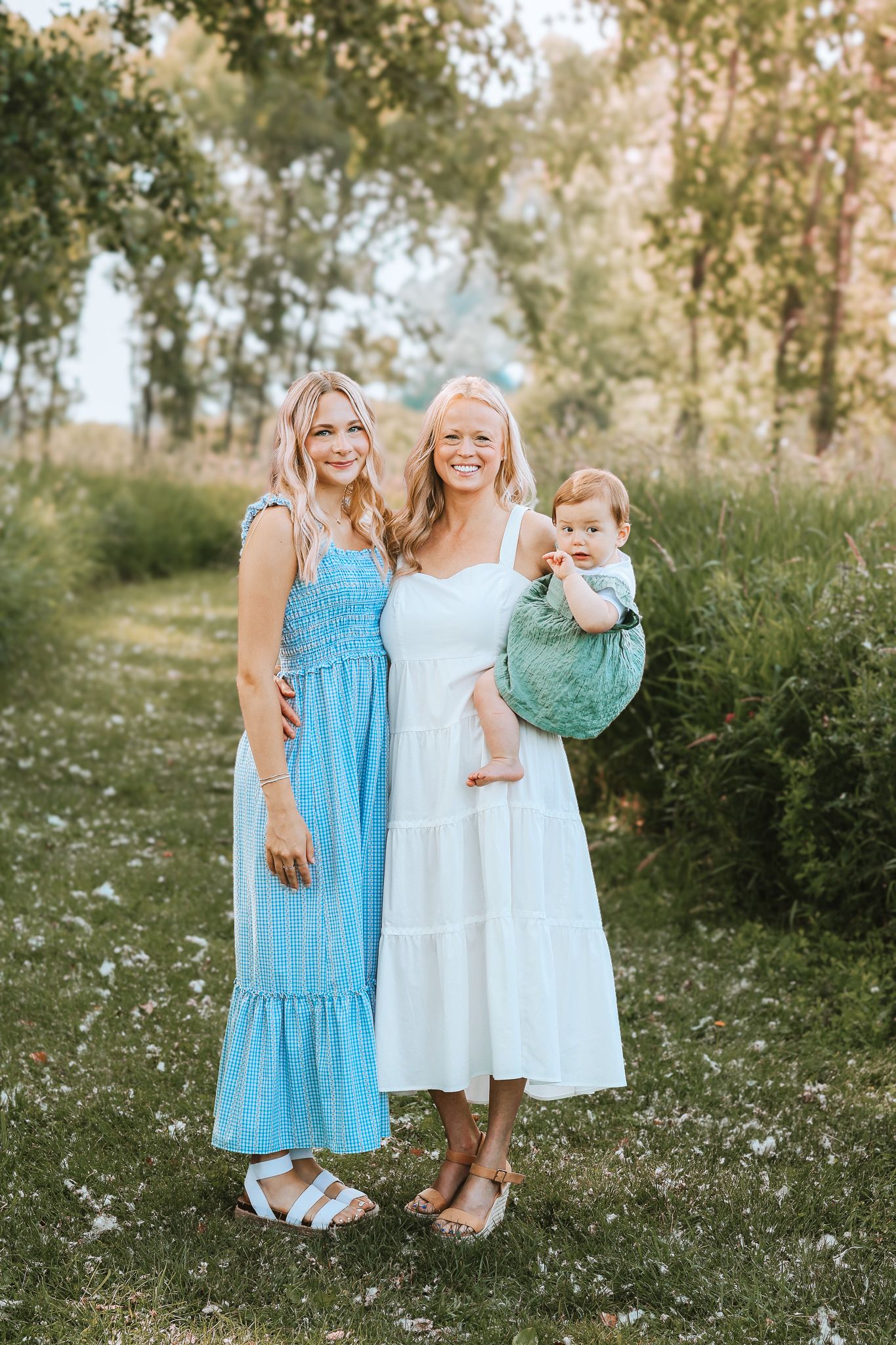 Two smiling blonde women in blue and white dresses holding a baby outdoors in a garden.