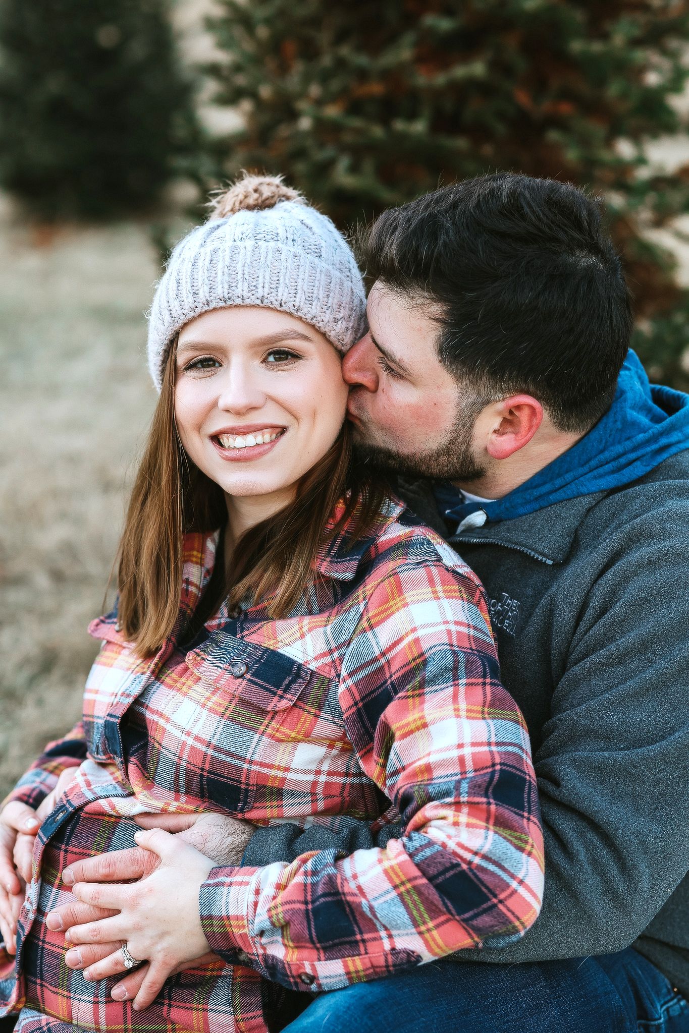 Man kissing smiling pregnant woman on cheek while embracing her outdoors near evergreen trees.