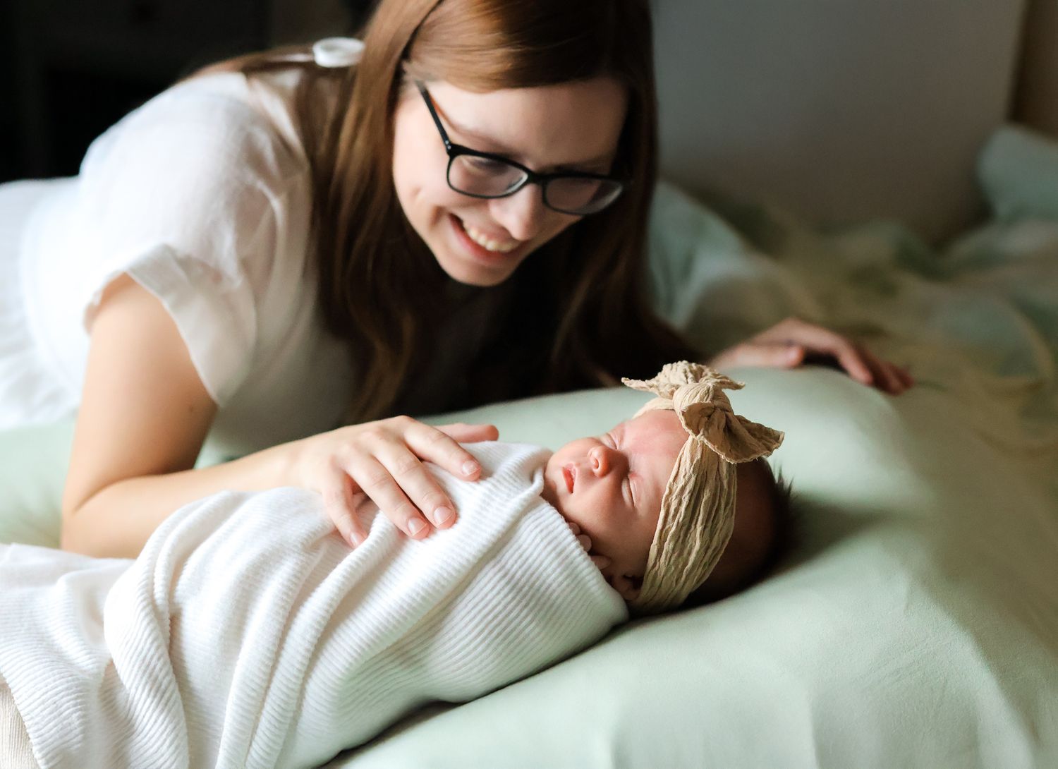 Mother with glasses smiling at her swaddled newborn baby girl wearing a floral headband on a bed.