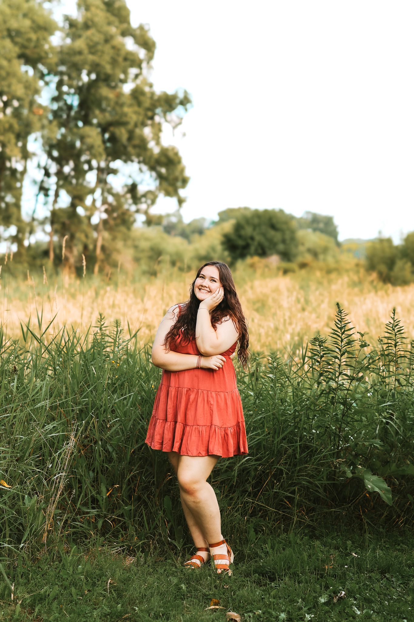 Smiling young woman in red dress standing in lush green field on sunny day.