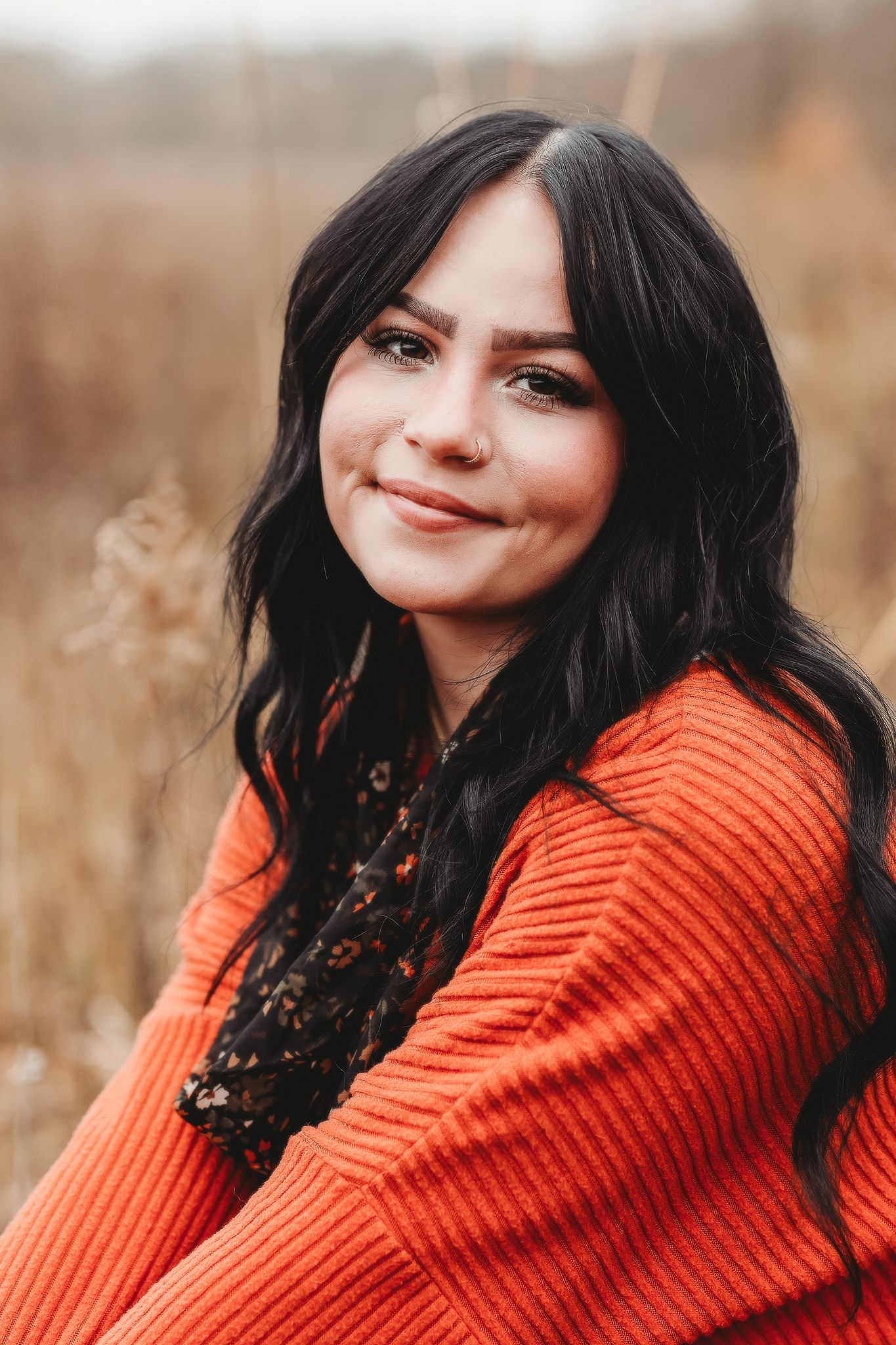 Close-up portrait of a dark-haired young woman in an orange sweater smiling in an autumn field.