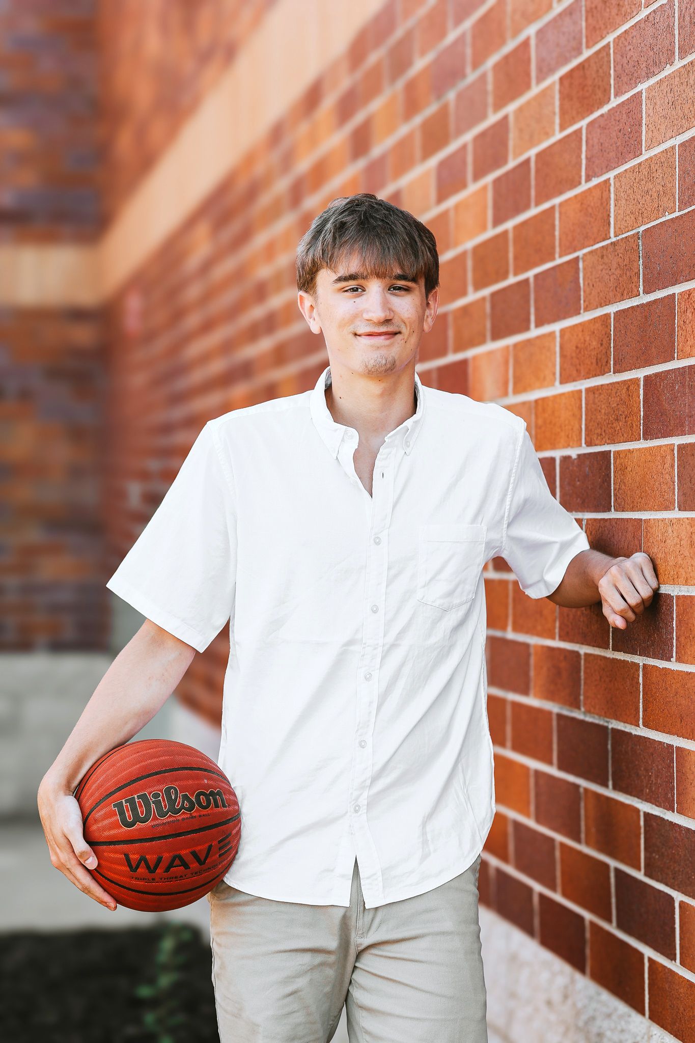 Smiling teenage boy in a white shirt holding a Wilson basketball while leaning against a red brick wall outdoors.