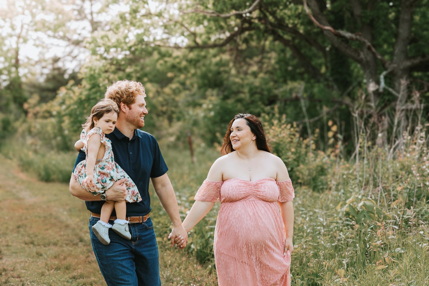 Expecting couple holding hands walking with toddler through a green outdoor meadow path.