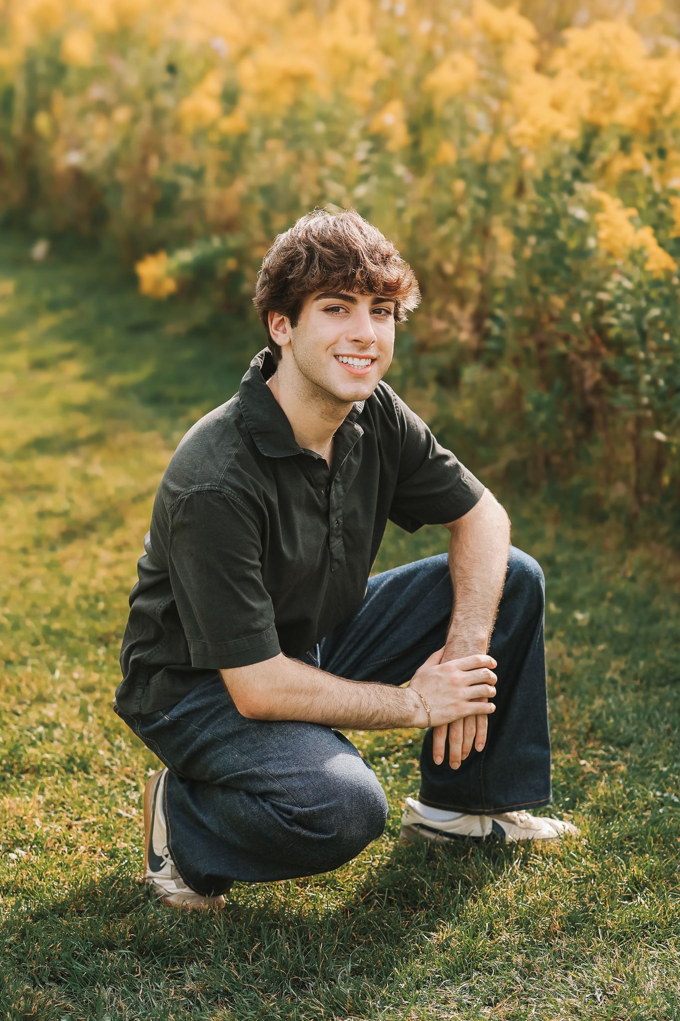 Smiling teen boy in dark polo shirt crouching on grass with golden autumn shrubs behind him.