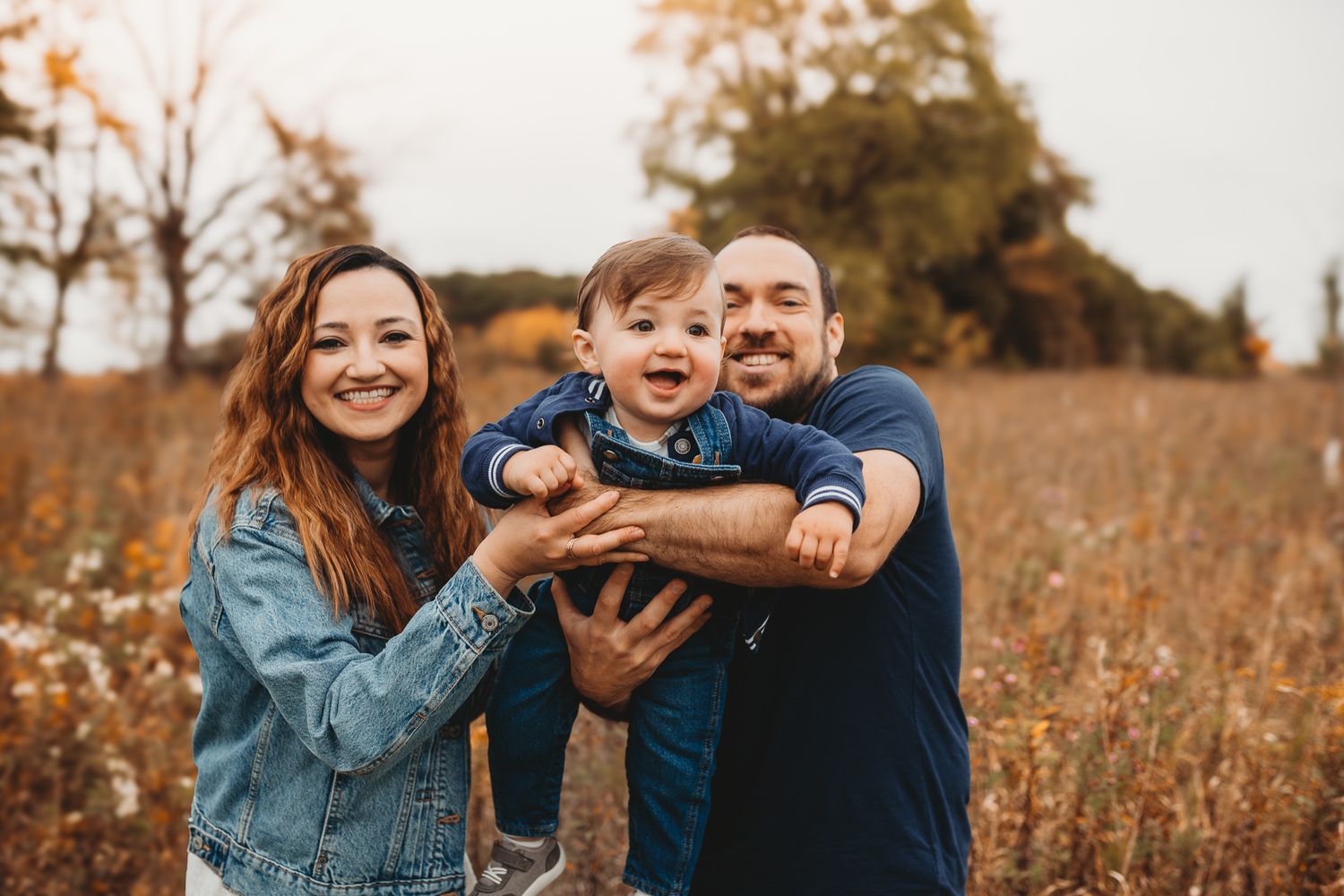 Happy family of three with parents holding laughing baby in autumn meadow outdoors.