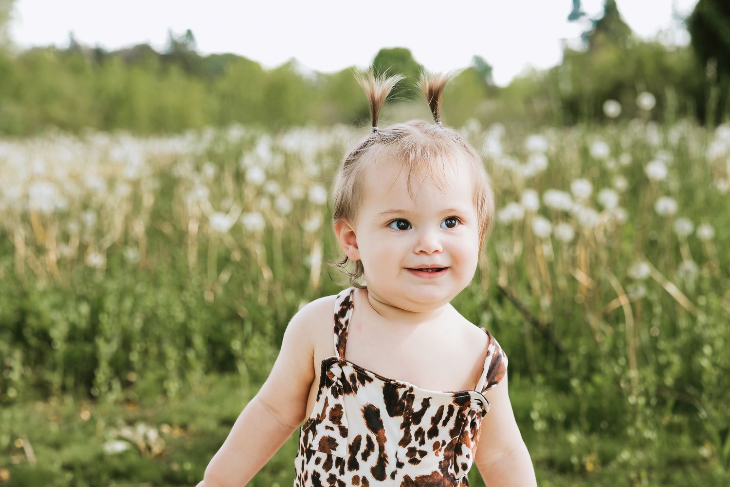Smiling baby girl in leopard print romper with tiny pigtails standing in a field of wildflowers.
