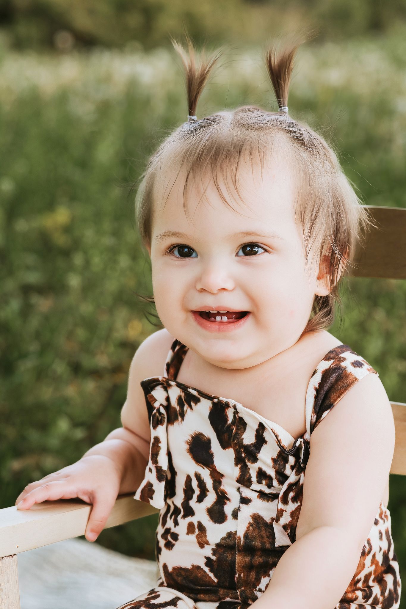 Happy smiling baby girl in leopard print romper leaning on wooden fence outdoors.