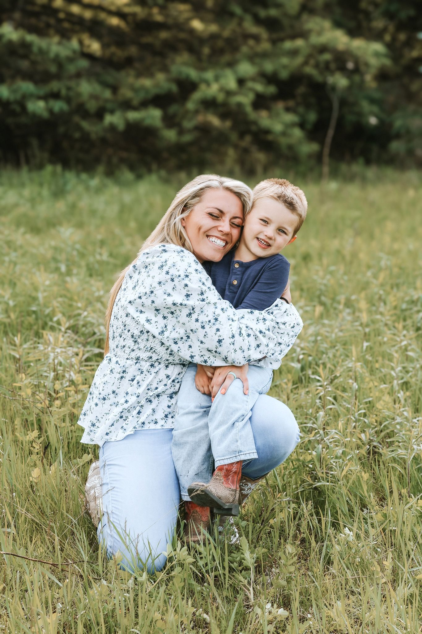 Blonde mother crouching in tall grass, hugging her young smiling son outdoors.