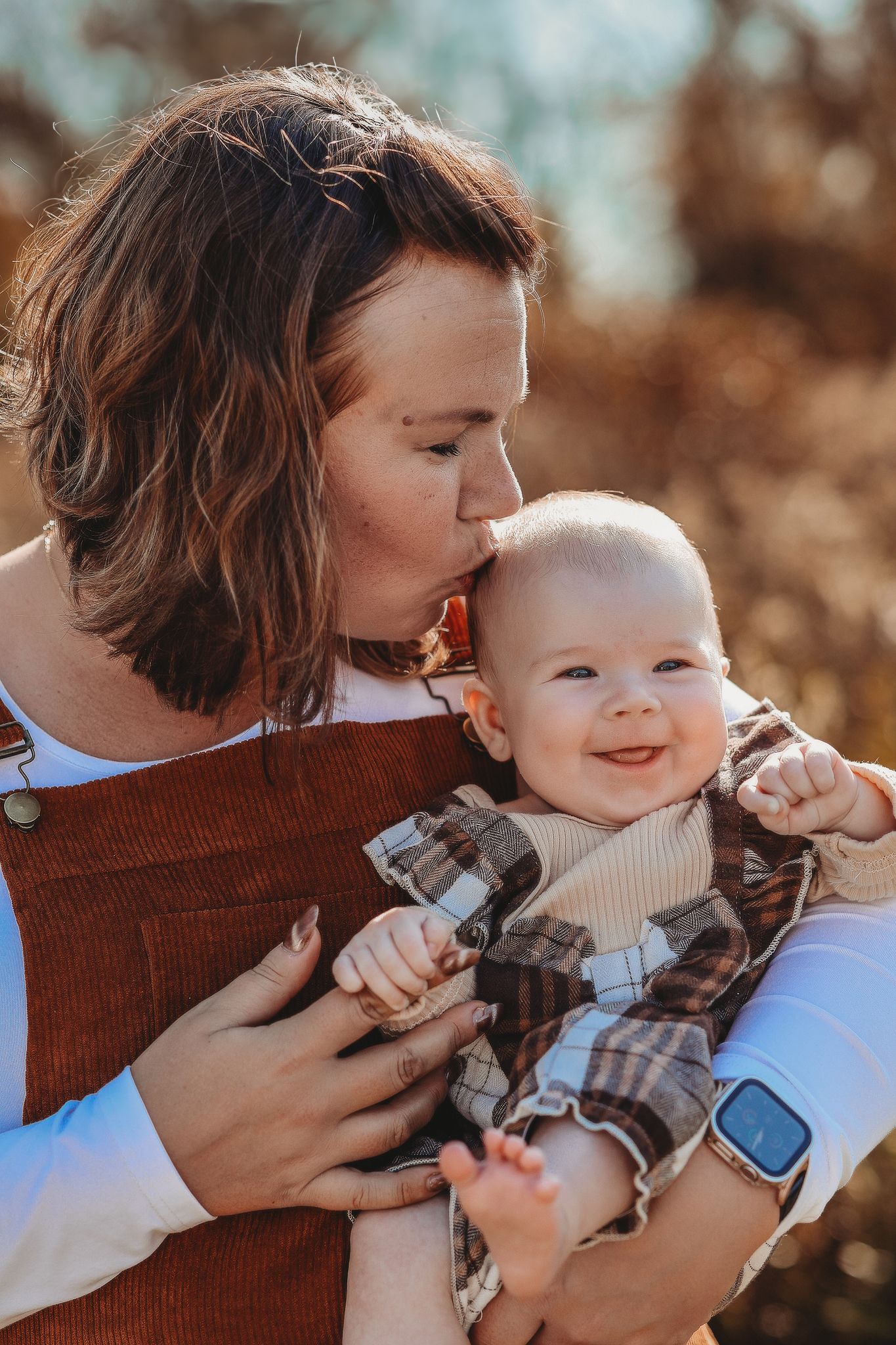 A mother tenderly kisses her smiling baby outdoors in warm autumn light.