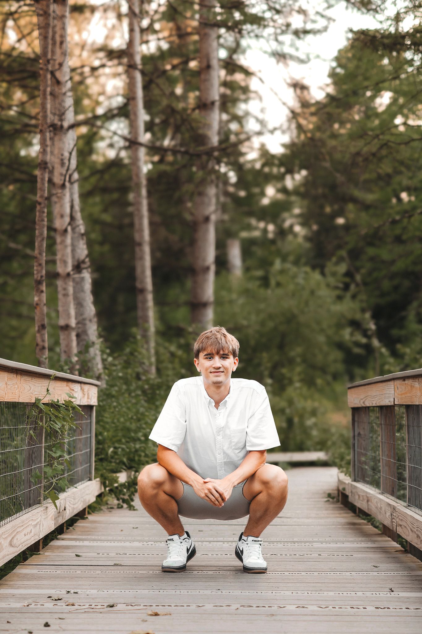 Young man in white shirt and gray shorts crouching on a wooden bridge surrounded by lush green forest.