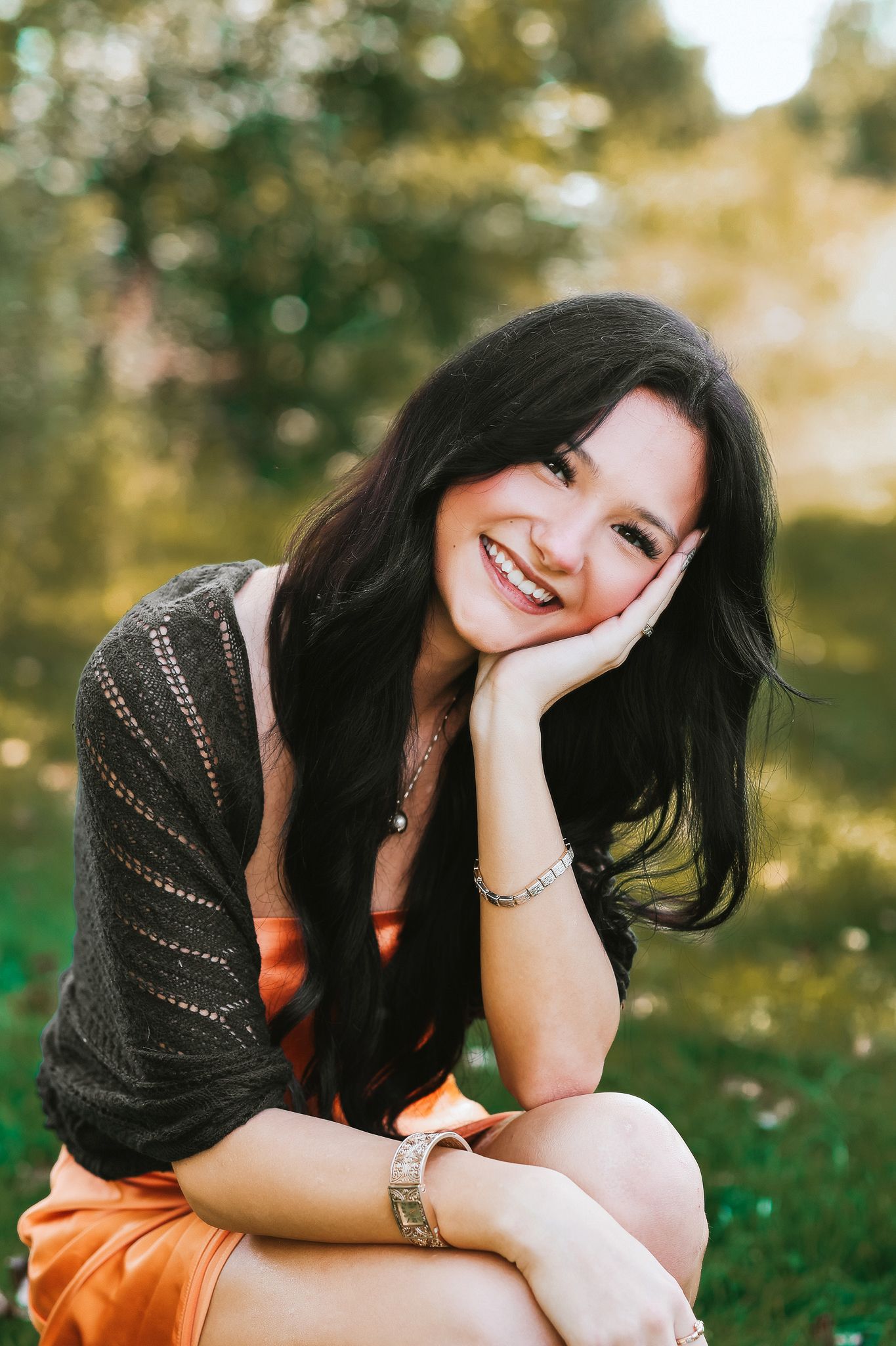 Happy young woman with long black hair sitting outdoors, resting cheek on hand, wearing orange skirt and dark cardigan.