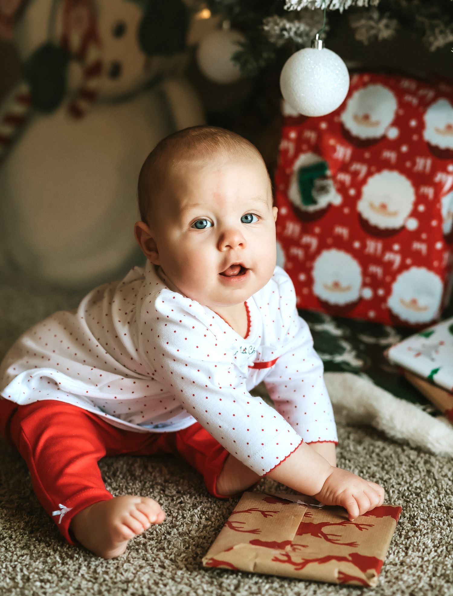 Baby girl in first Christmas outfit sitting near wrapped gifts under a Christmas tree.