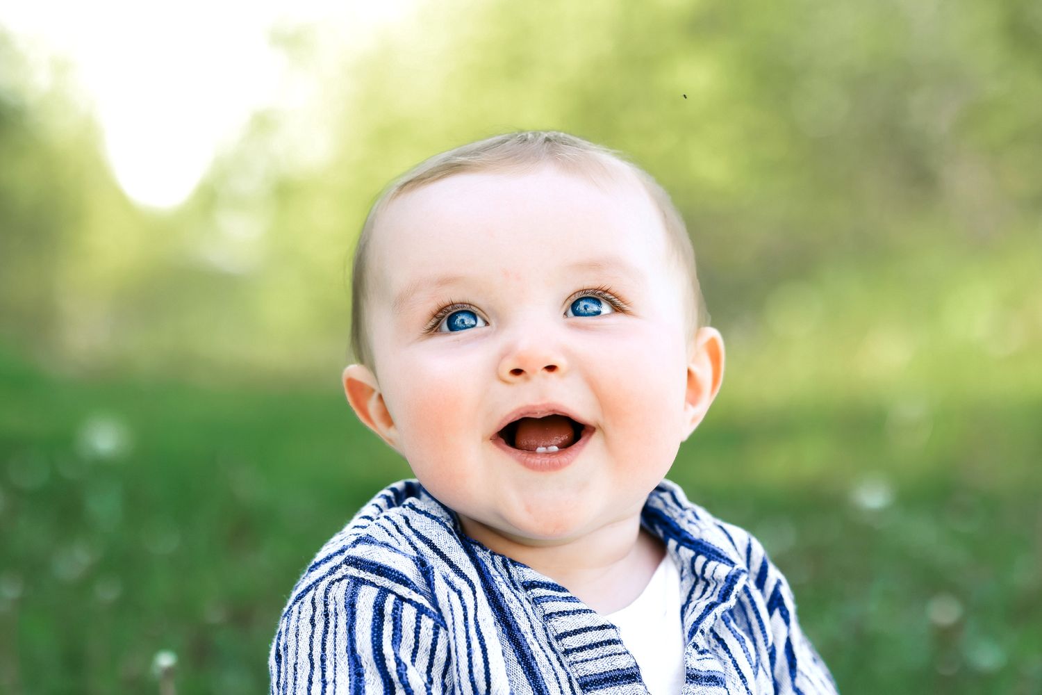 Cheerful blue-eyed baby boy in striped blue jacket laughing joyfully in sunny green outdoor setting.