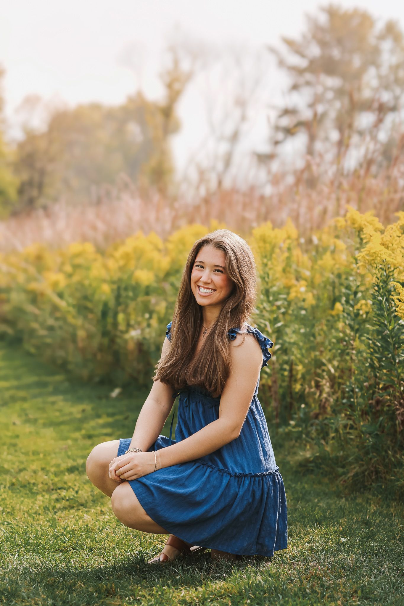 Happy young woman in blue sundress posing outdoors among vibrant autumn wildflowers.