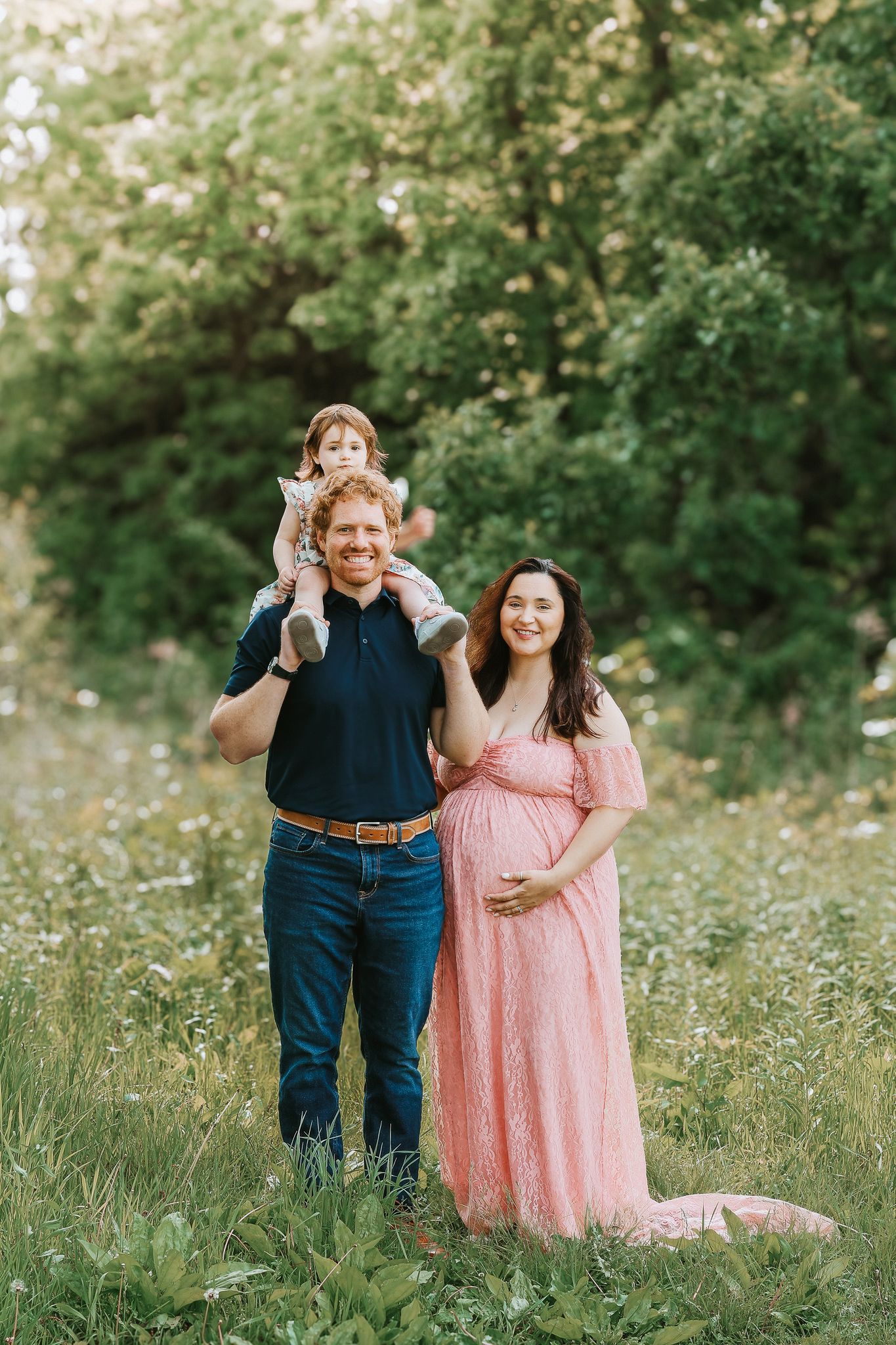 Pregnant woman in pink lace gown with husband and toddler in a wildflower meadow.