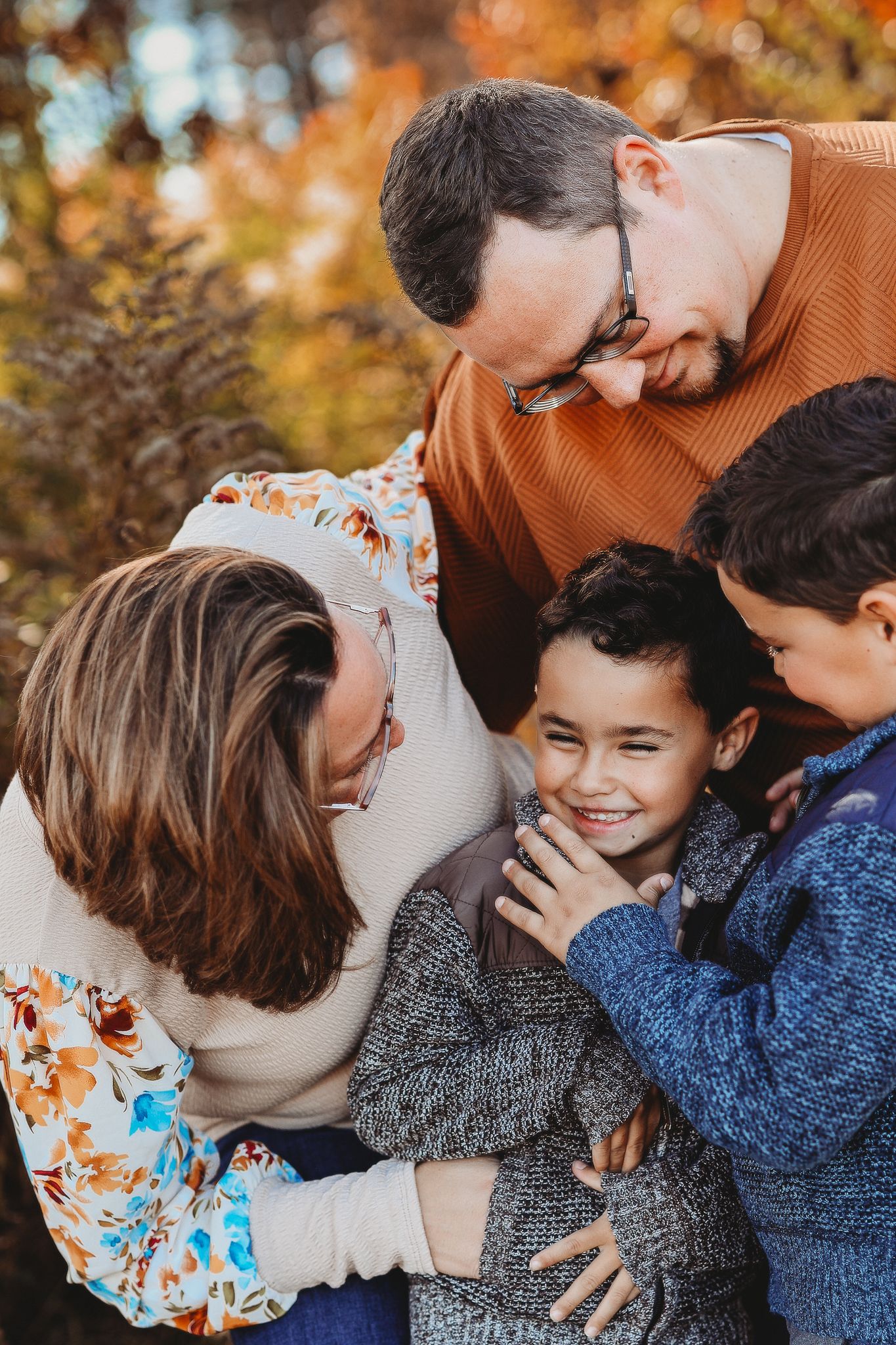 Two loving parents lean in smiling at their laughing child in autumn outdoor setting.