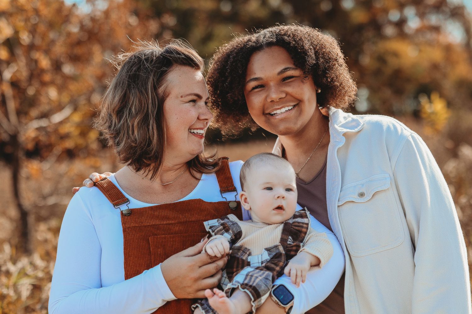 Two women smile warmly while holding an adorable baby outdoors in fall setting.