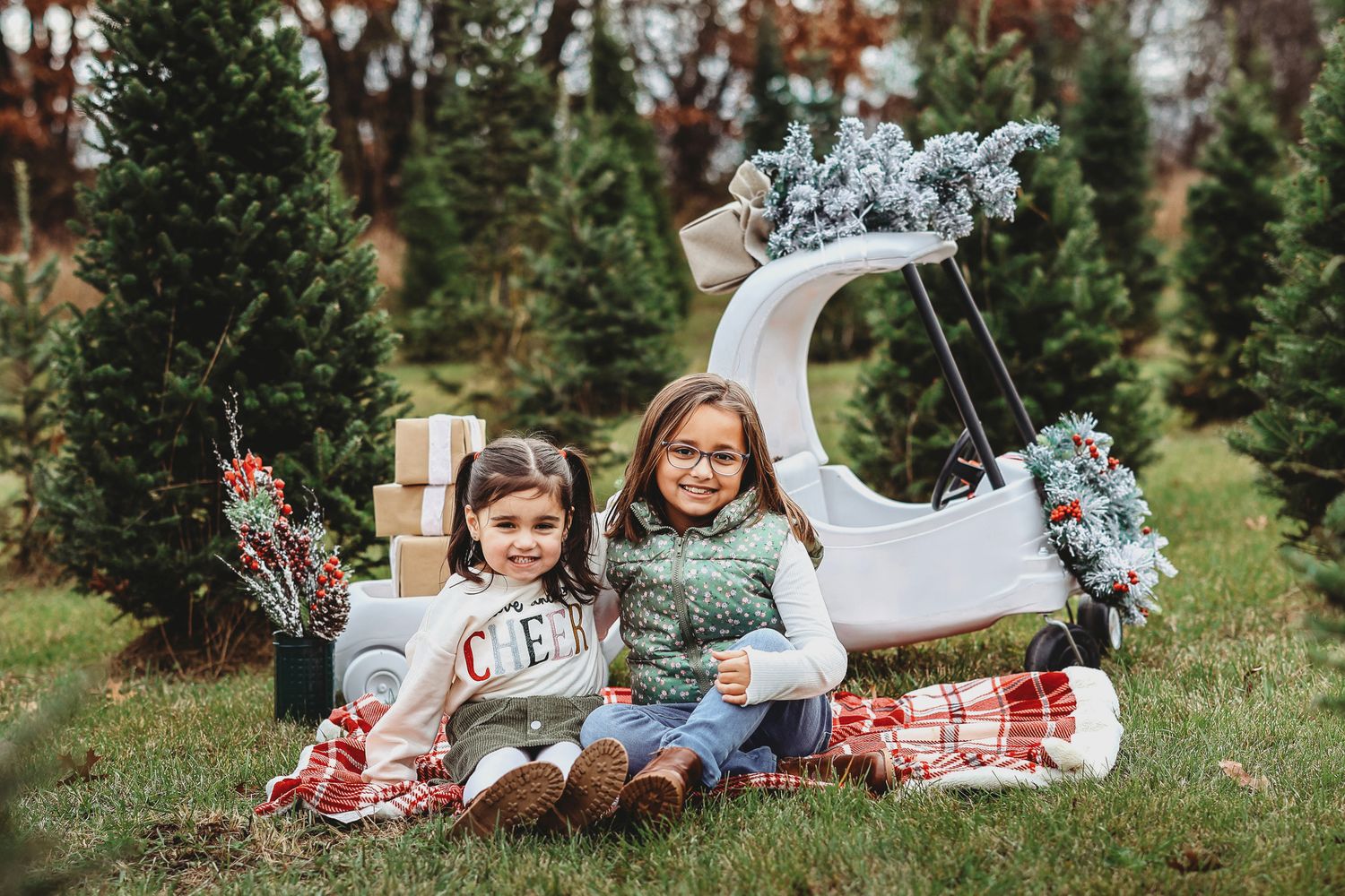 Two smiling girls pose together on a plaid blanket with a decorated sleigh at a Christmas tree farm.