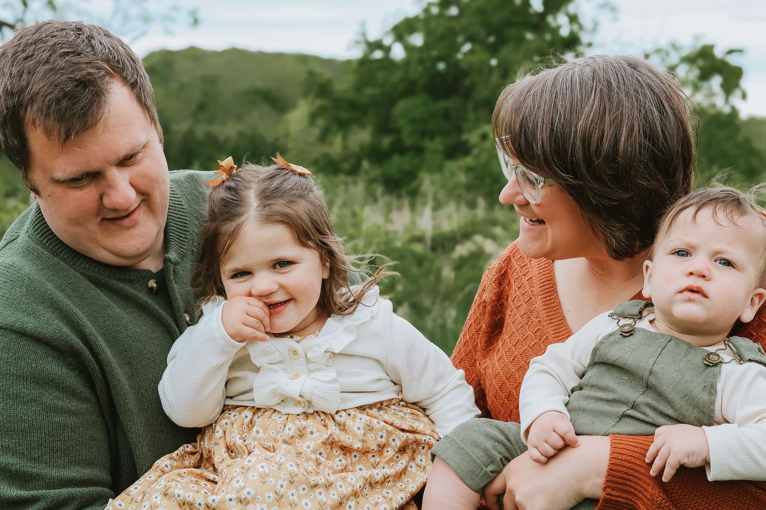 Two parents smile while holding their toddler girl and baby boy outdoors in nature.