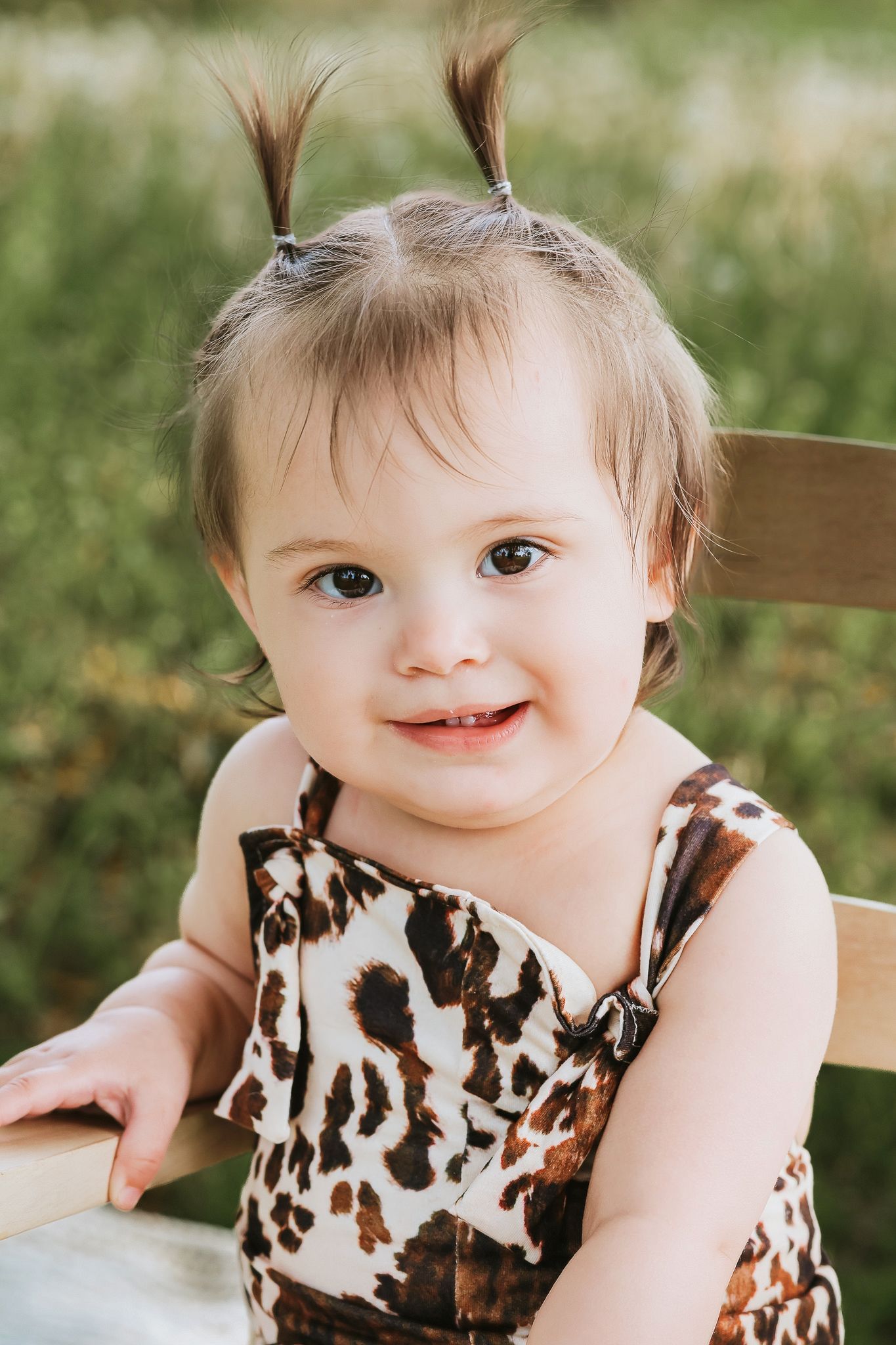 Close-up of a happy baby girl in leopard print dress sitting in a wooden chair smiling outdoors.