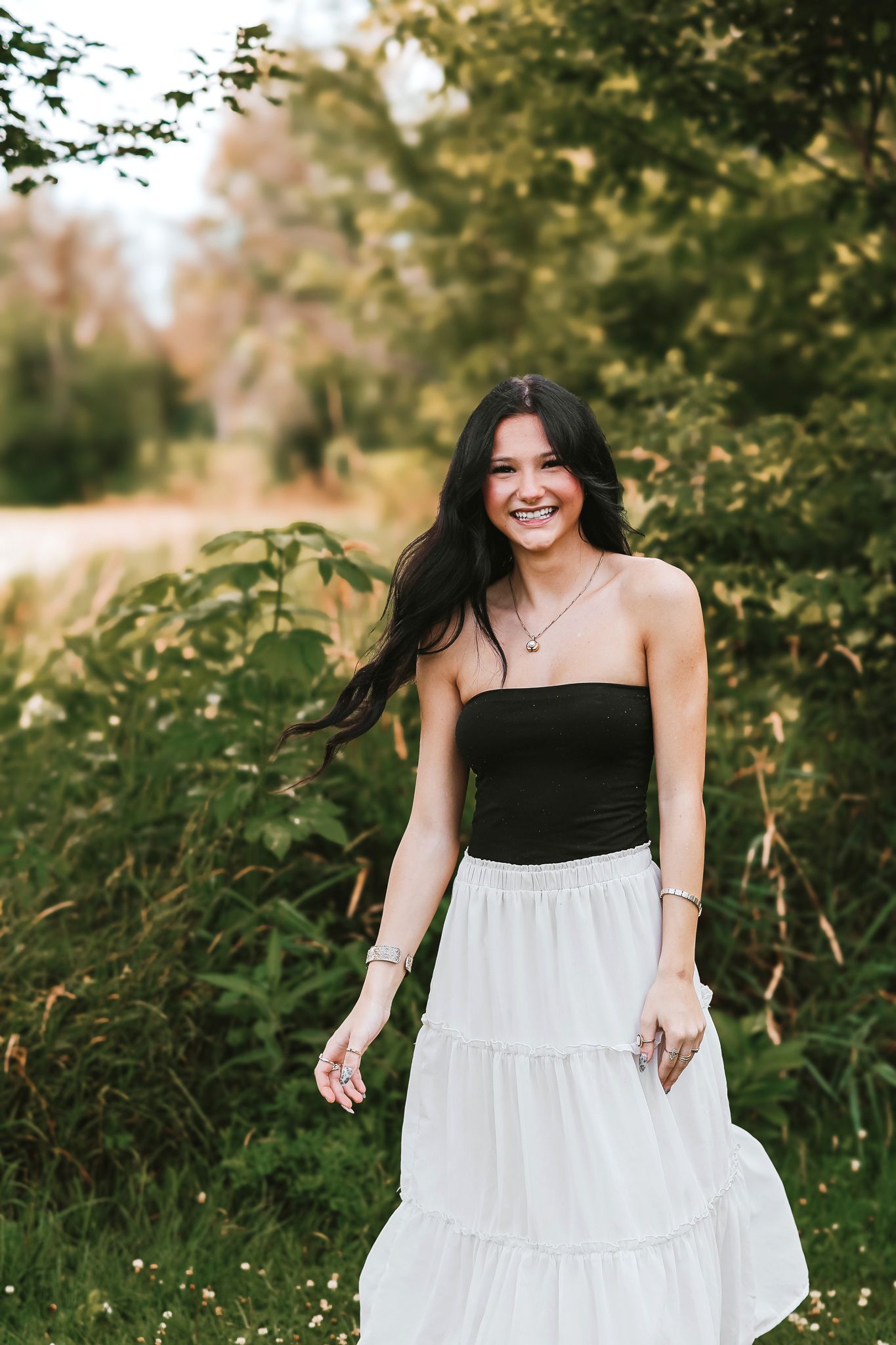Joyful young woman with long dark hair in a black tube top and white tiered skirt walking along a green path.