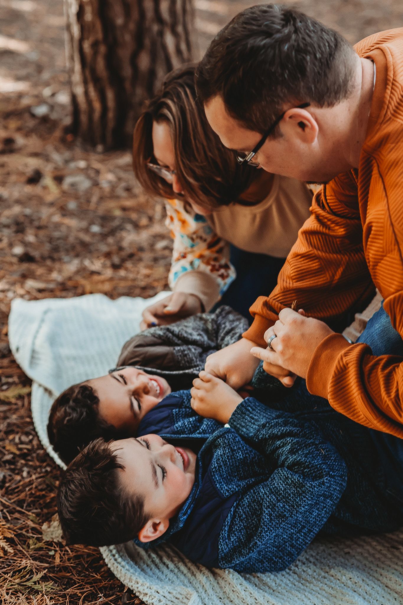 Parents tickling and playing with their laughing young son on a blanket in autumn woods.