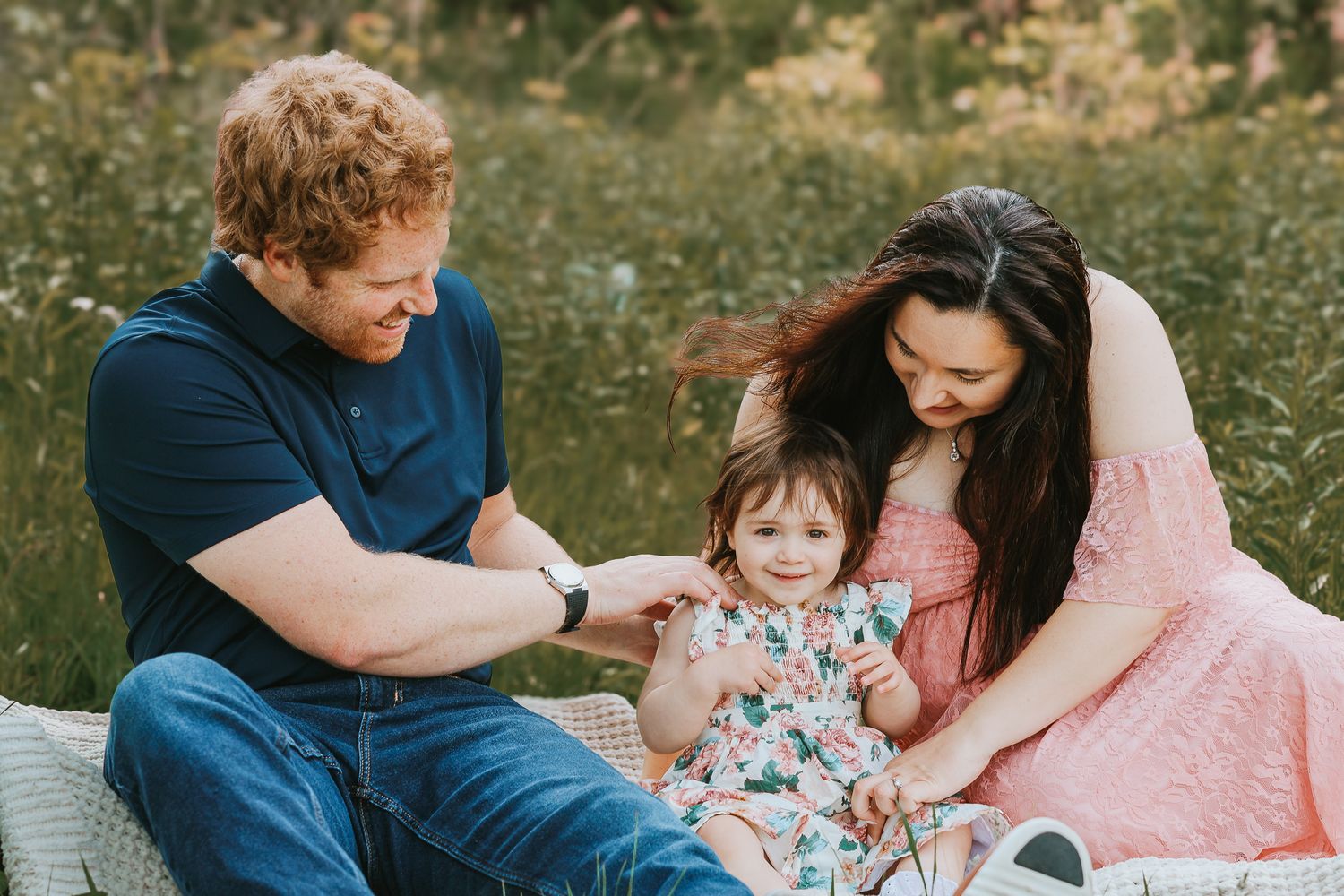 Young family with redhead dad and dark-haired mom in pink dress playing with toddler in wildflower field.