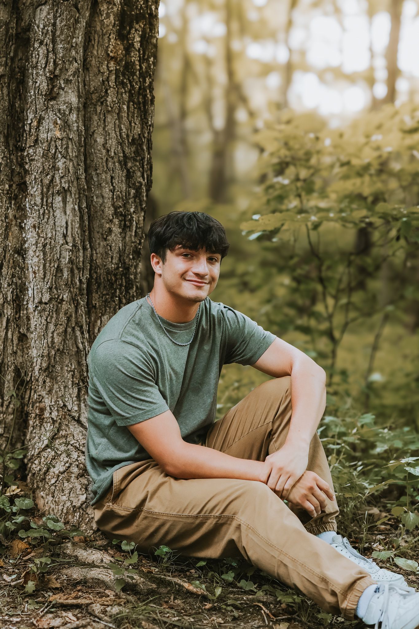 Young man in gray tee and khakis sitting against tree trunk in forest setting.